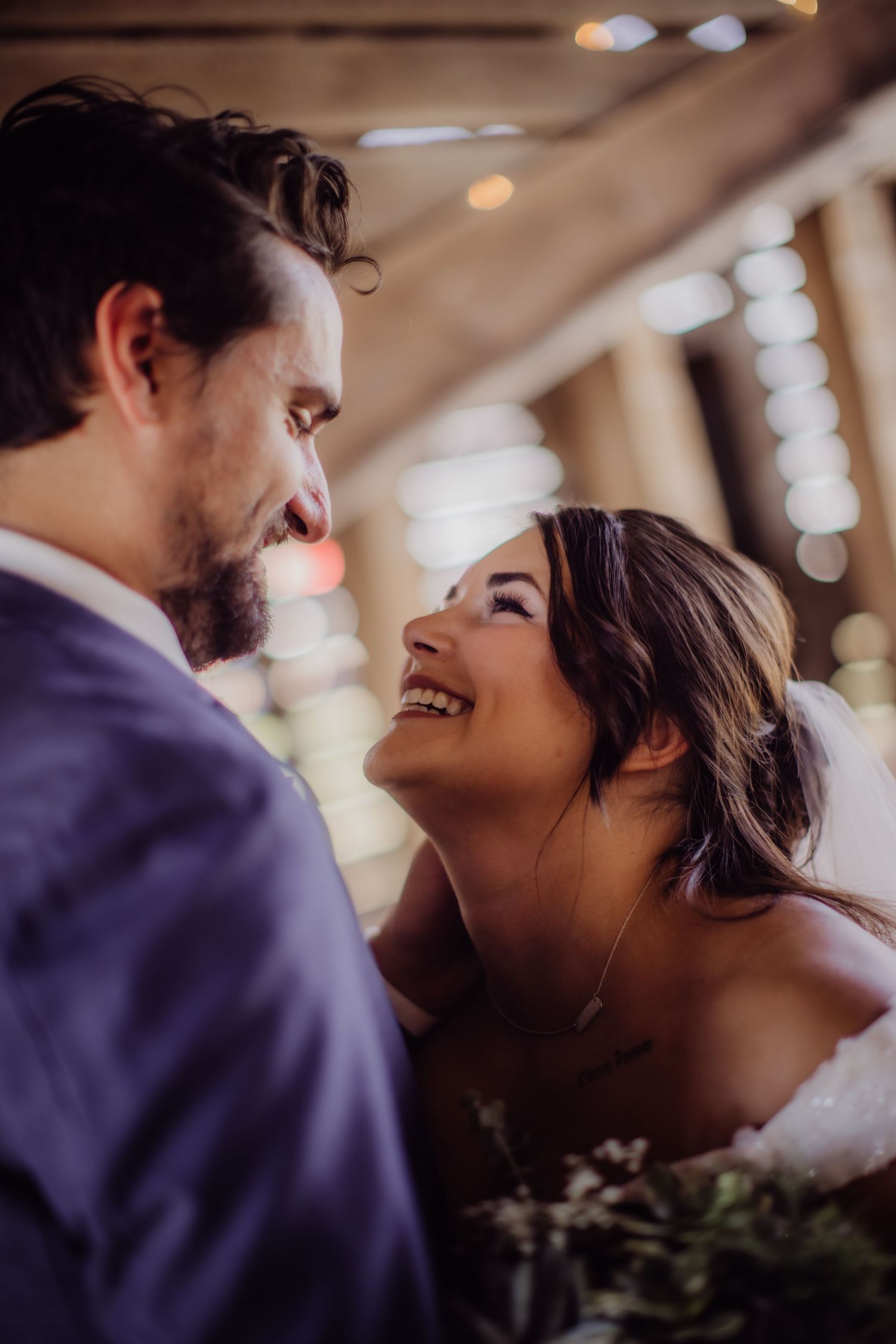 Candid wedding portrait showing loving couple under twinkling lights in rustic setting.