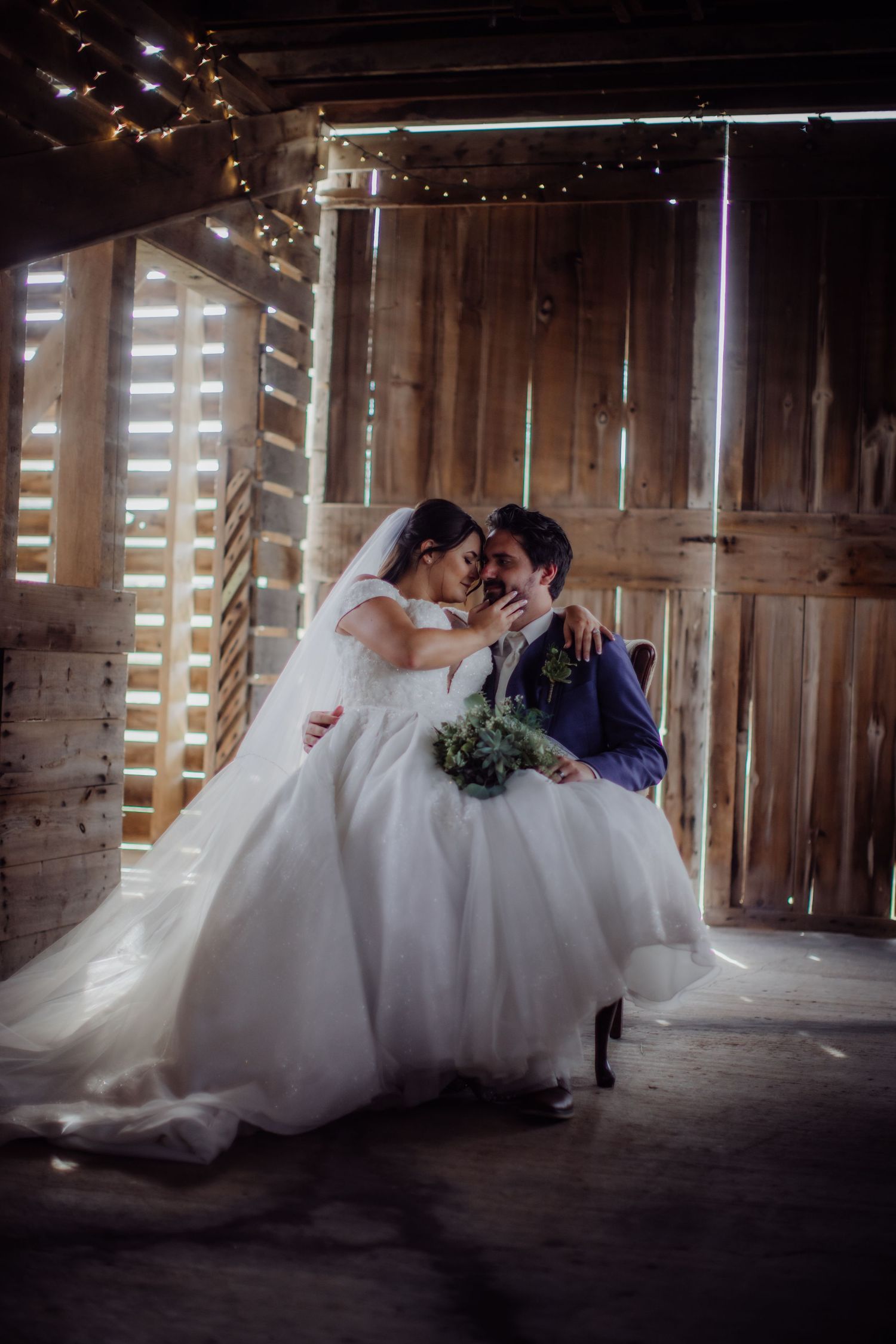 Intimate wedding portrait in a rustic barn with twinkling lights overhead.