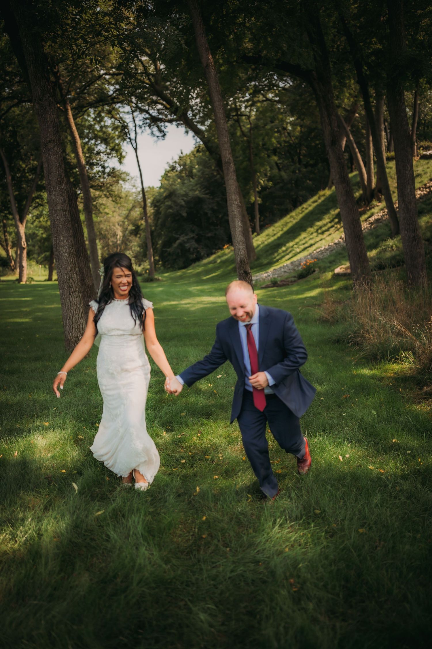 A couple runs playfully through a grassy park setting during a wedding photoshoot.