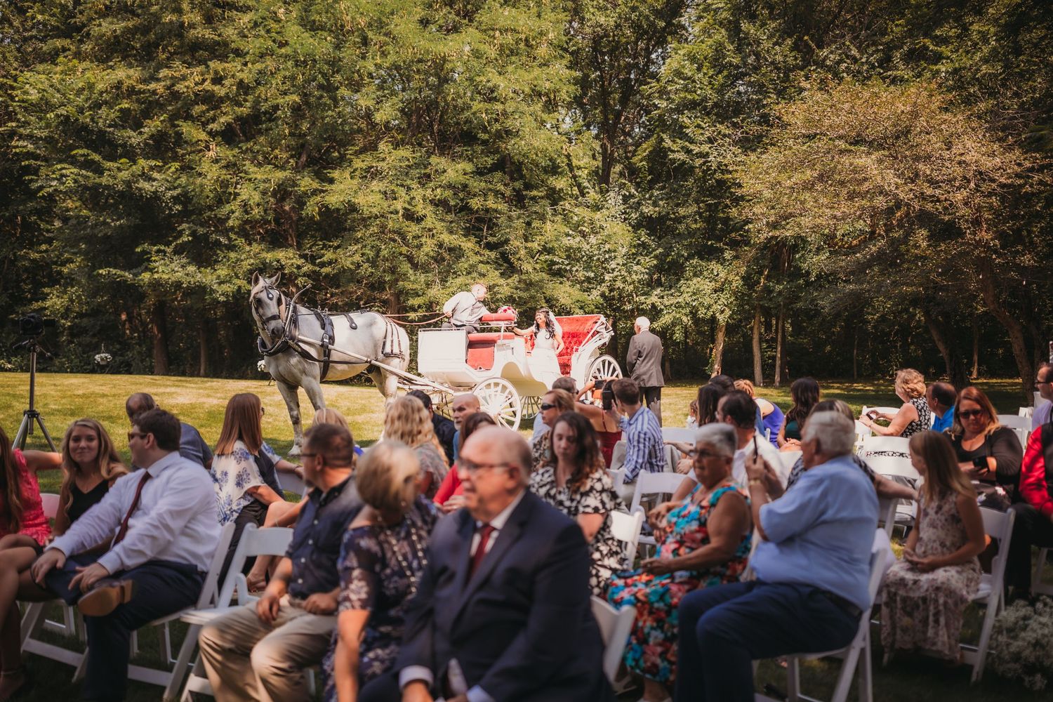Guests seated in white chairs at an outdoor wedding ceremony with trees in background.