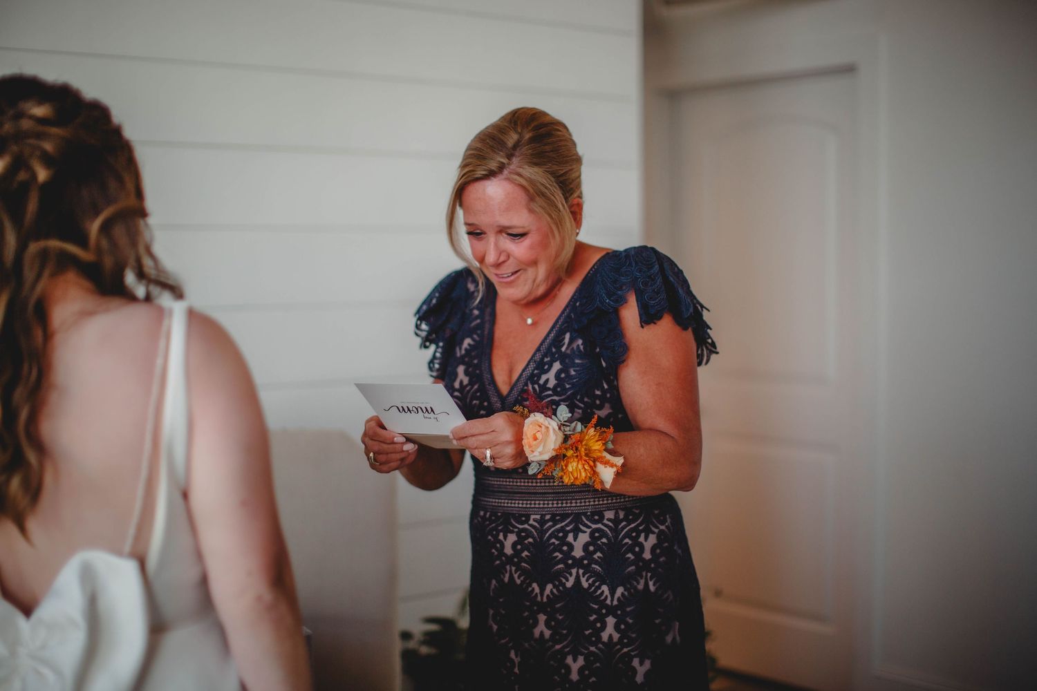 Someone in a navy lace dress reads from a card while standing with someone in a white wedding gown.