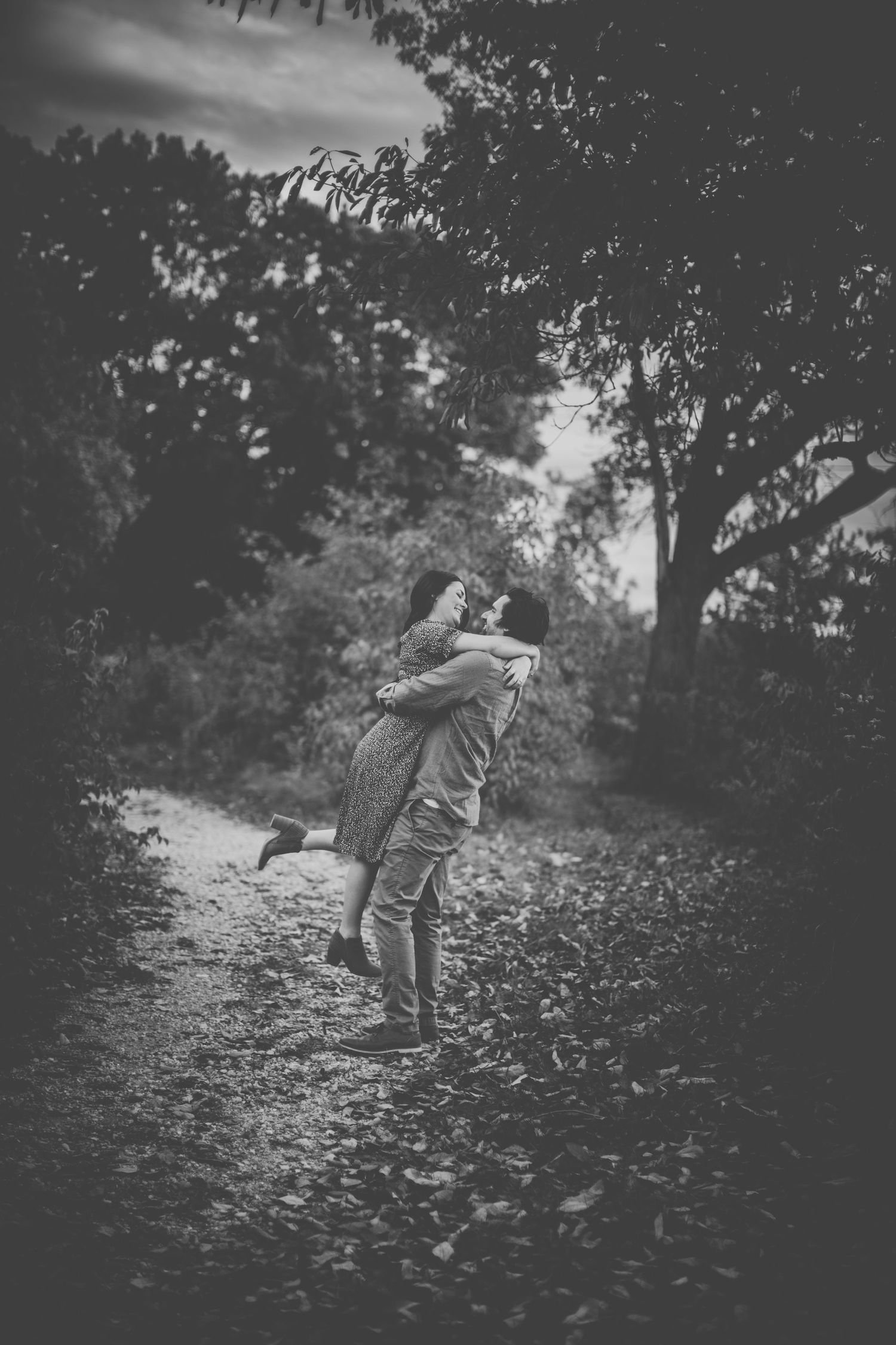 Black and white photo captures romantic embrace on wooded autumn path.