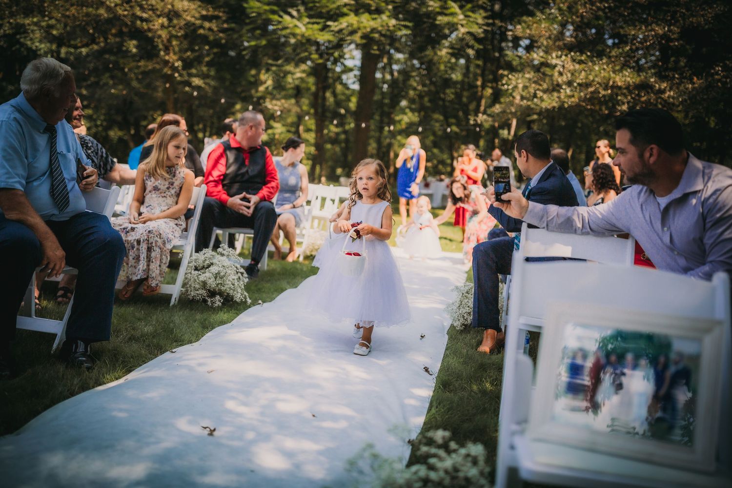 A child in white dress walks down an outdoor aisle as wedding guests look on from both sides.