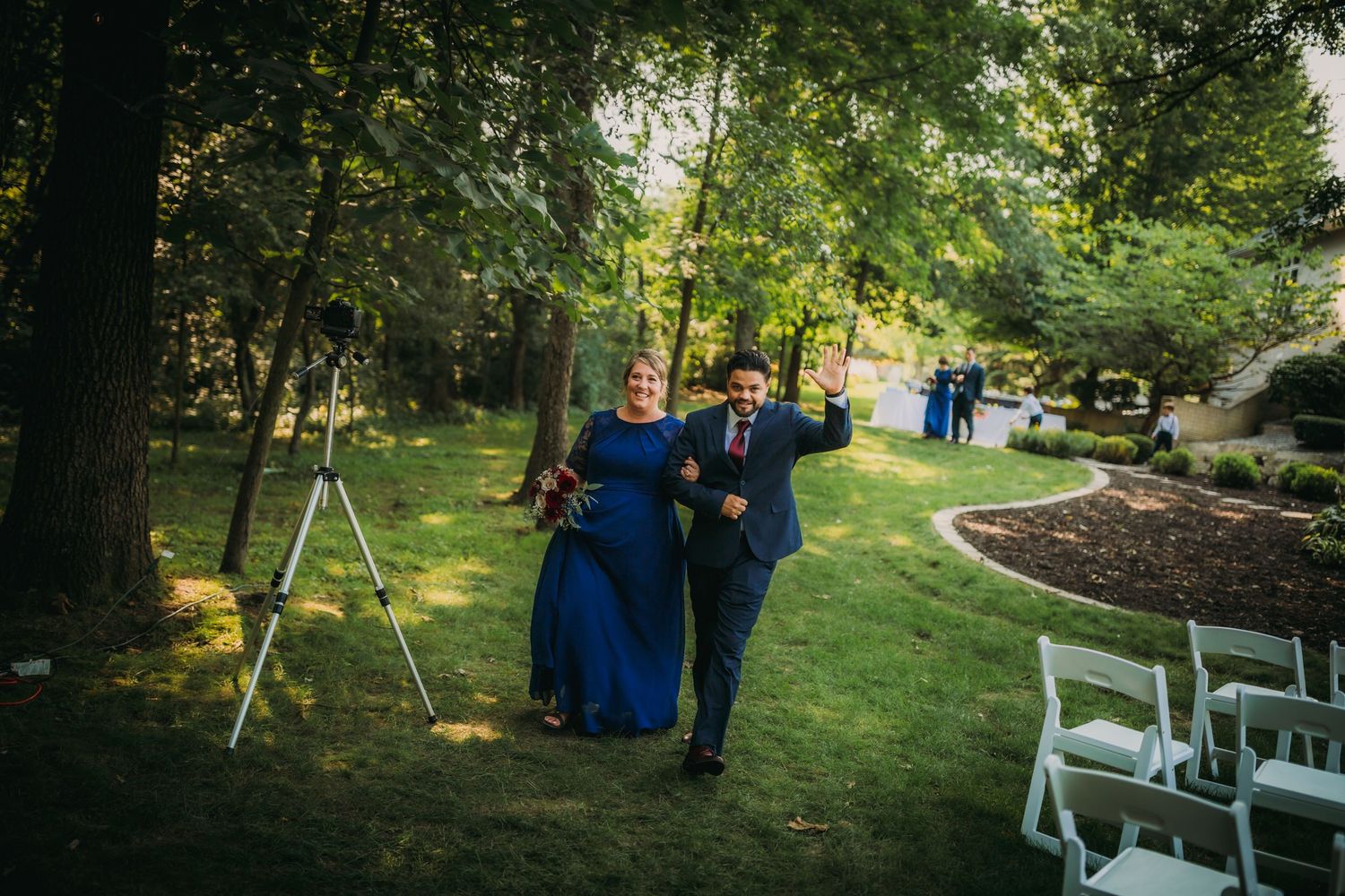 Wedding attendants stroll down an outdoor aisle lined with white chairs and trees.