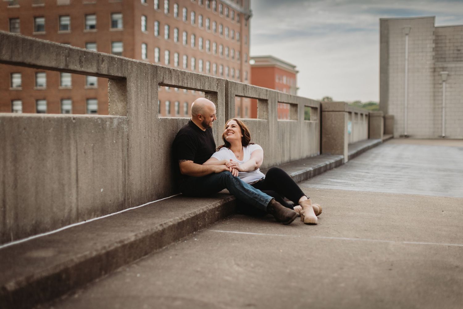 A couple lounges together on a concrete wall with red brick buildings in the background.