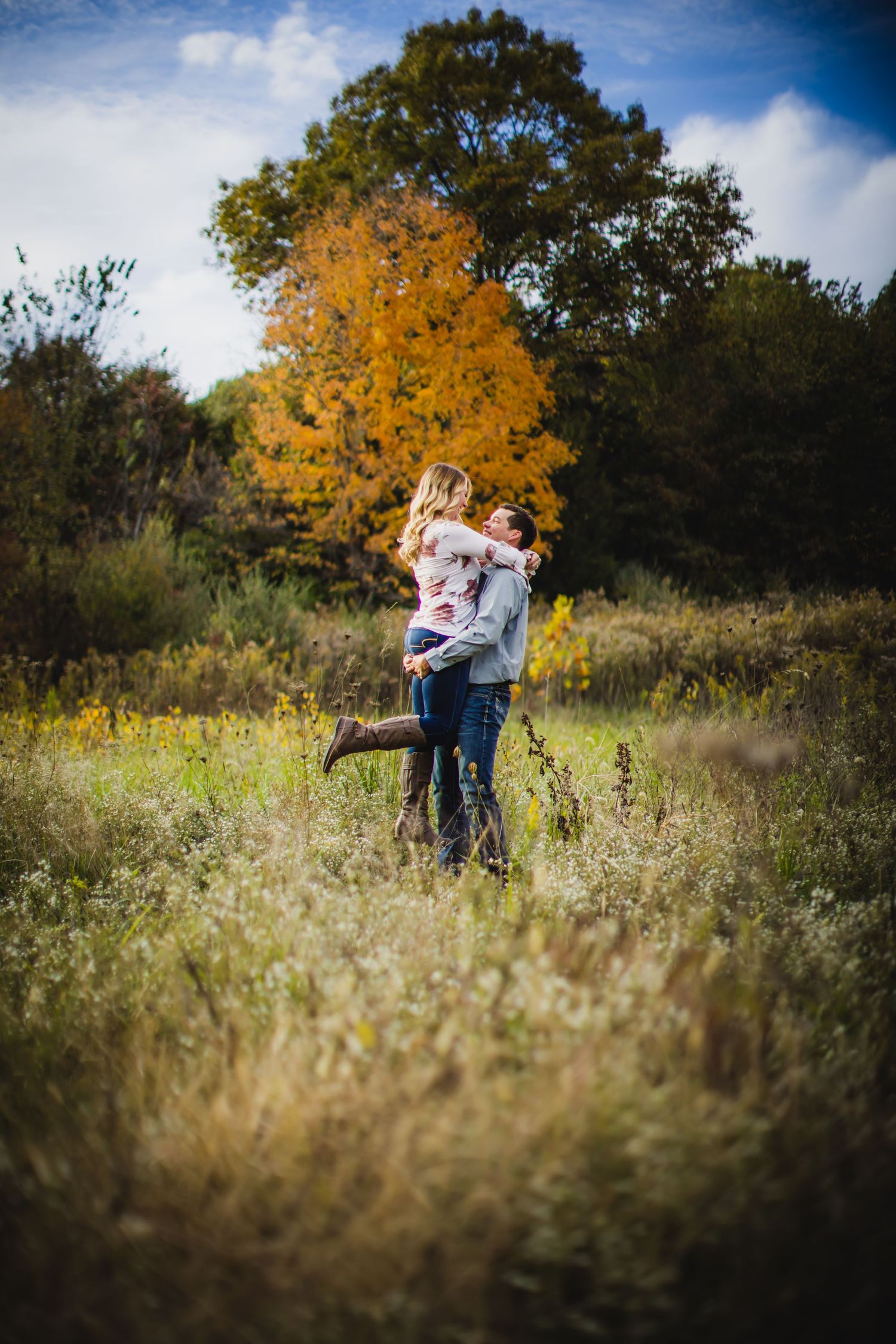 An autumn engagement photo featuring a couple embracing in a field with golden trees behind them.