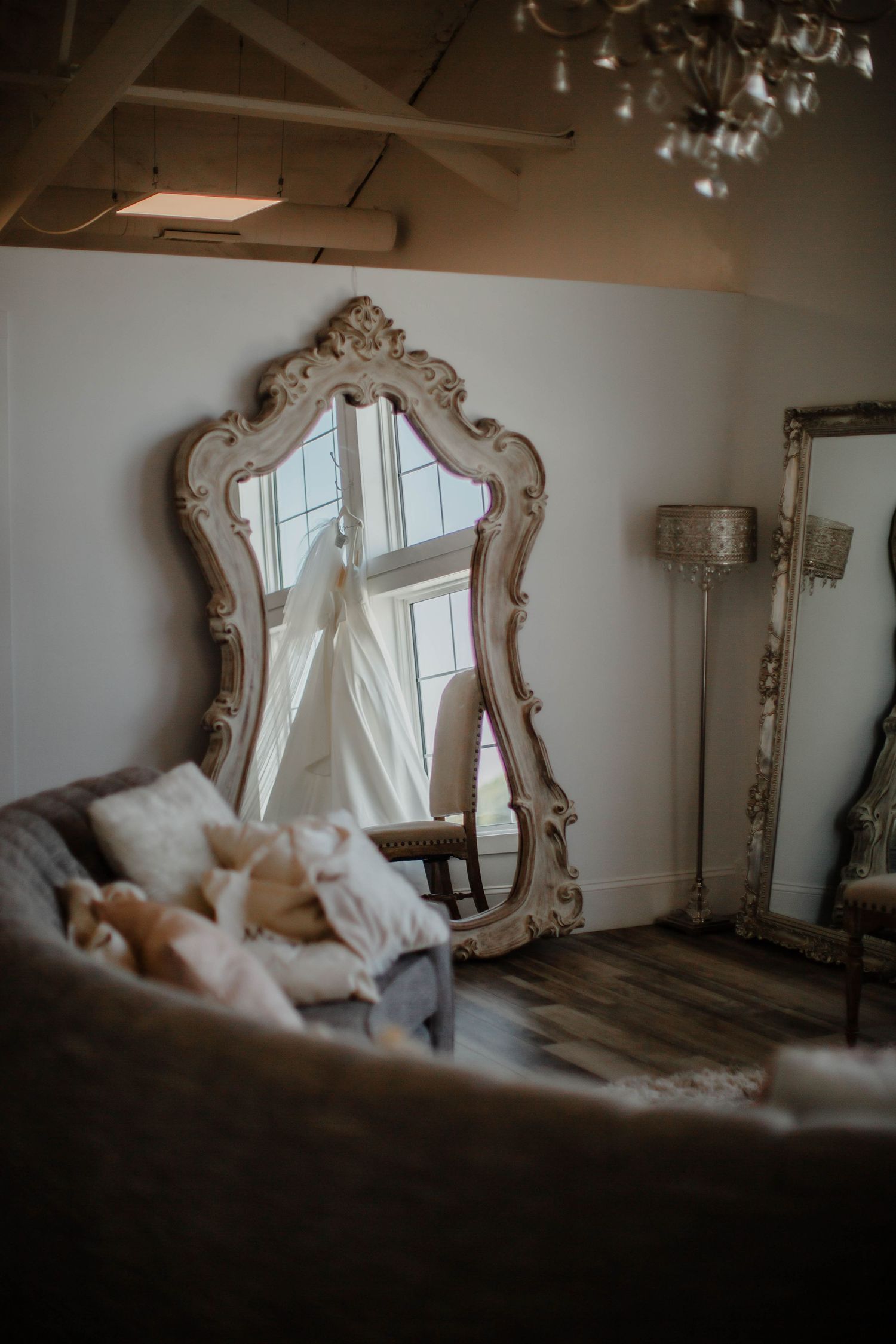 Elegant white wedding dress hanging near ornate vintage mirrors in a sunlit room.