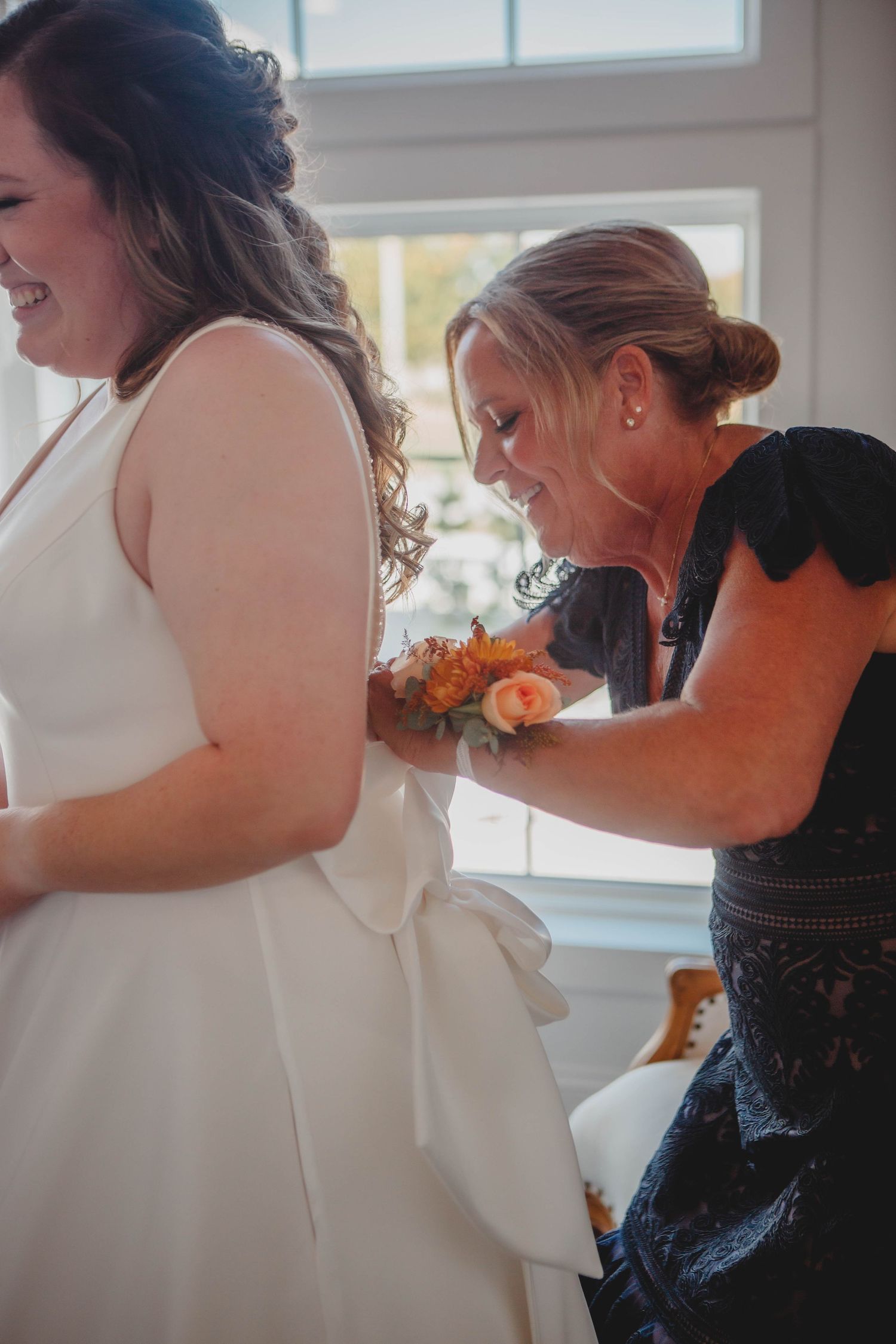 A woman in a white wedding dress being assisted with final touches by someone wearing a navy lace dress.