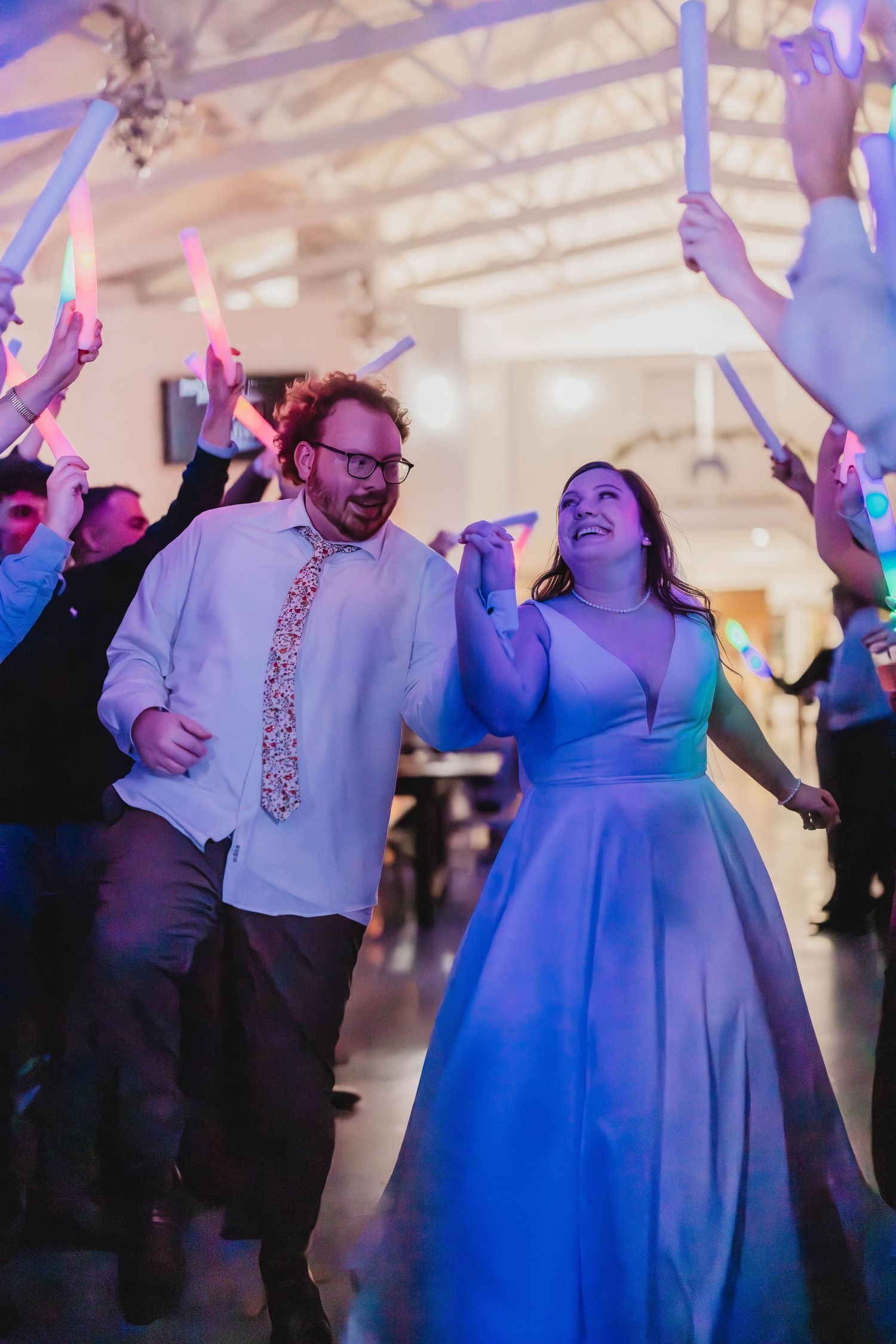 Joyful couple laughs while dancing under raised glow sticks at their wedding reception.