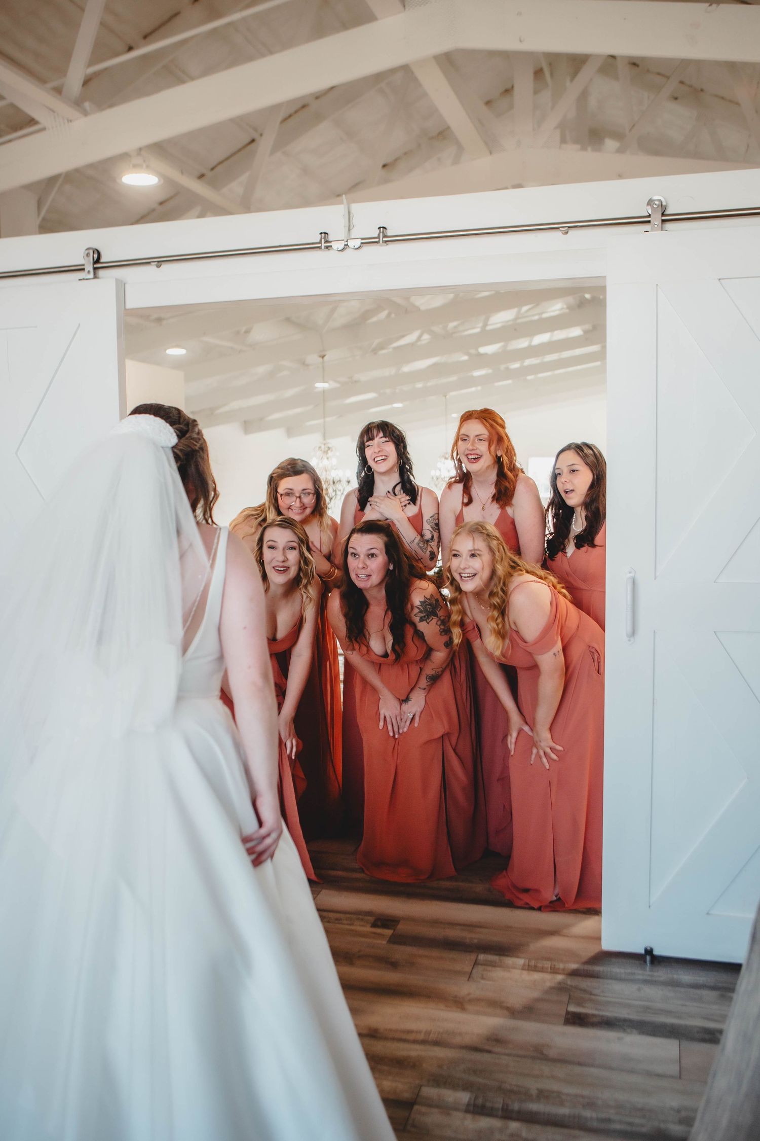 Wedding reveal moment as bridesmaids in coral dresses react to seeing bride through doorway.