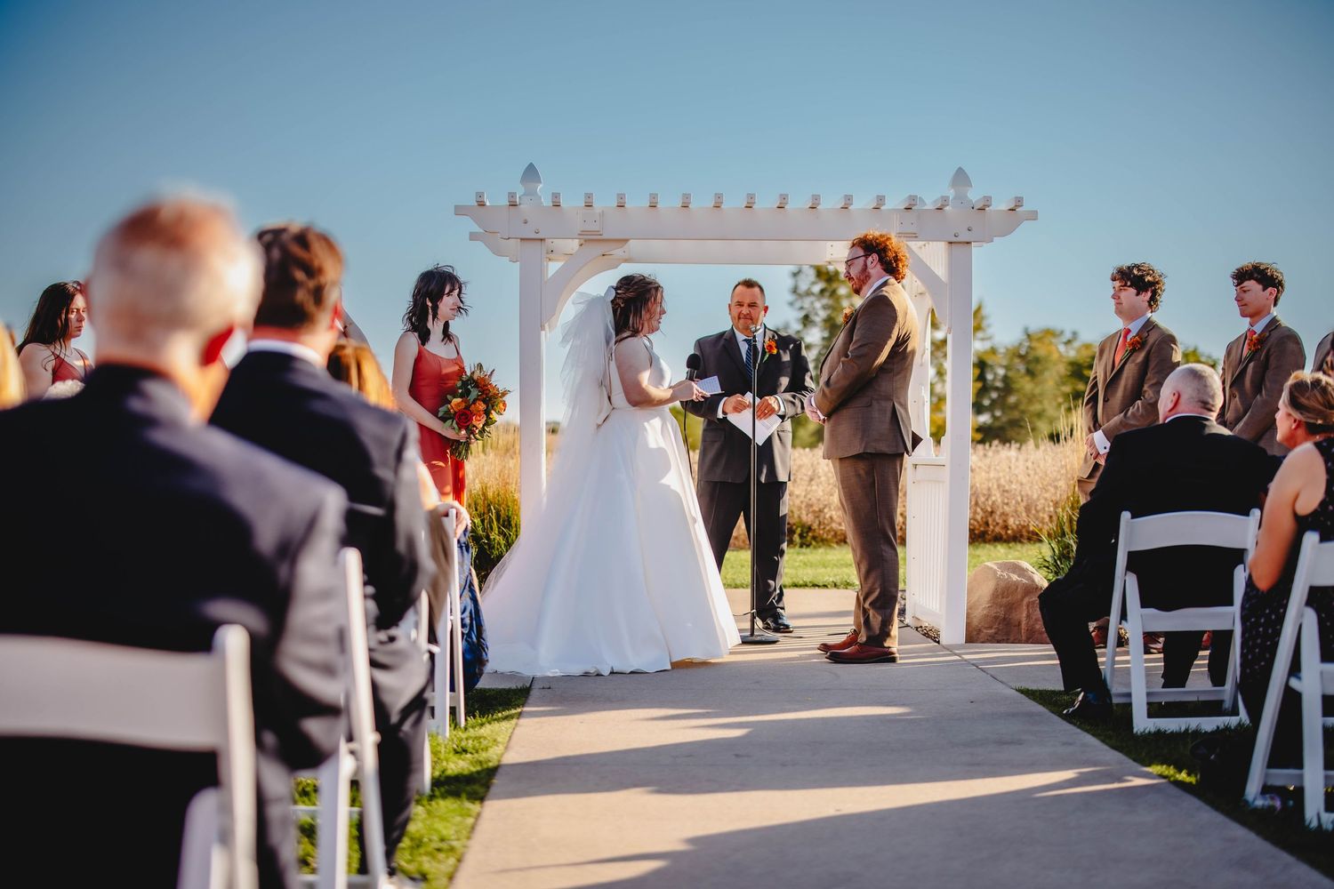 Wedding ceremony in progress under a white pergola with guests seated on white chairs.