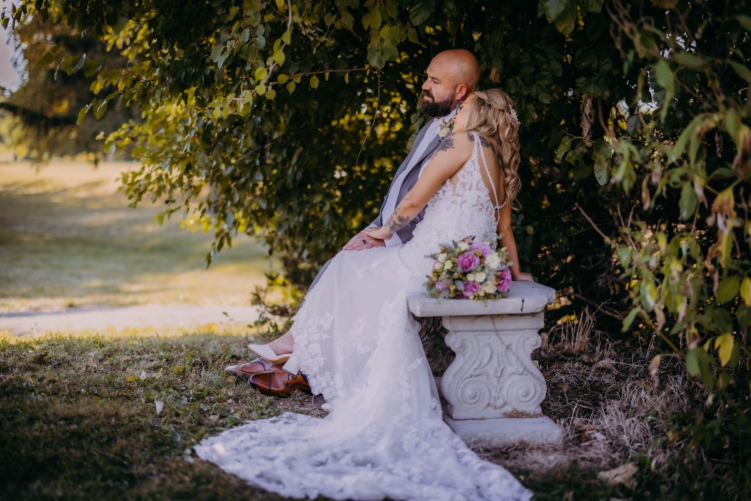A artistic wedding photo showing a couple sitting on a stone bench surrounded by lush foliage.