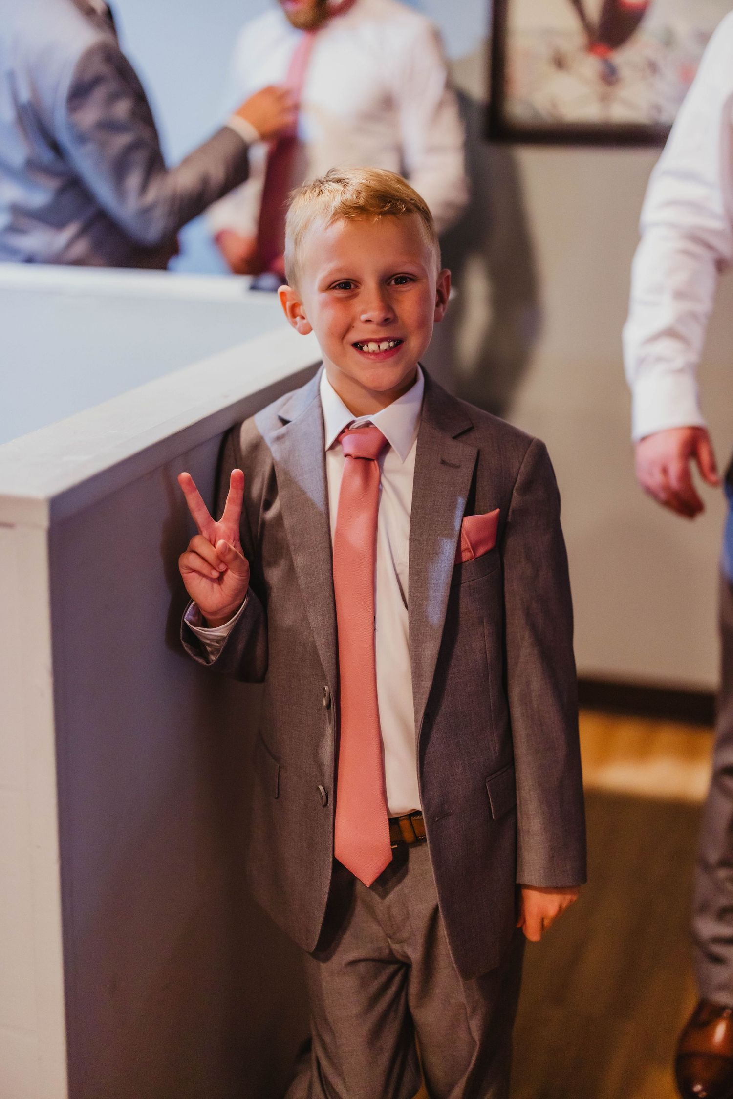 Young attendee in a grey suit and salmon tie making peace sign at formal event.