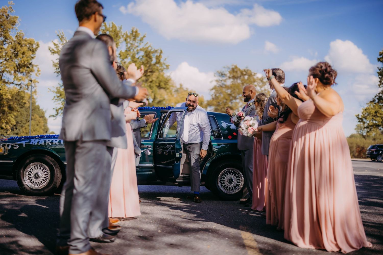 Wedding party gathers around a vintage car on a sunny day wearing gray suits and blush pink dresses.