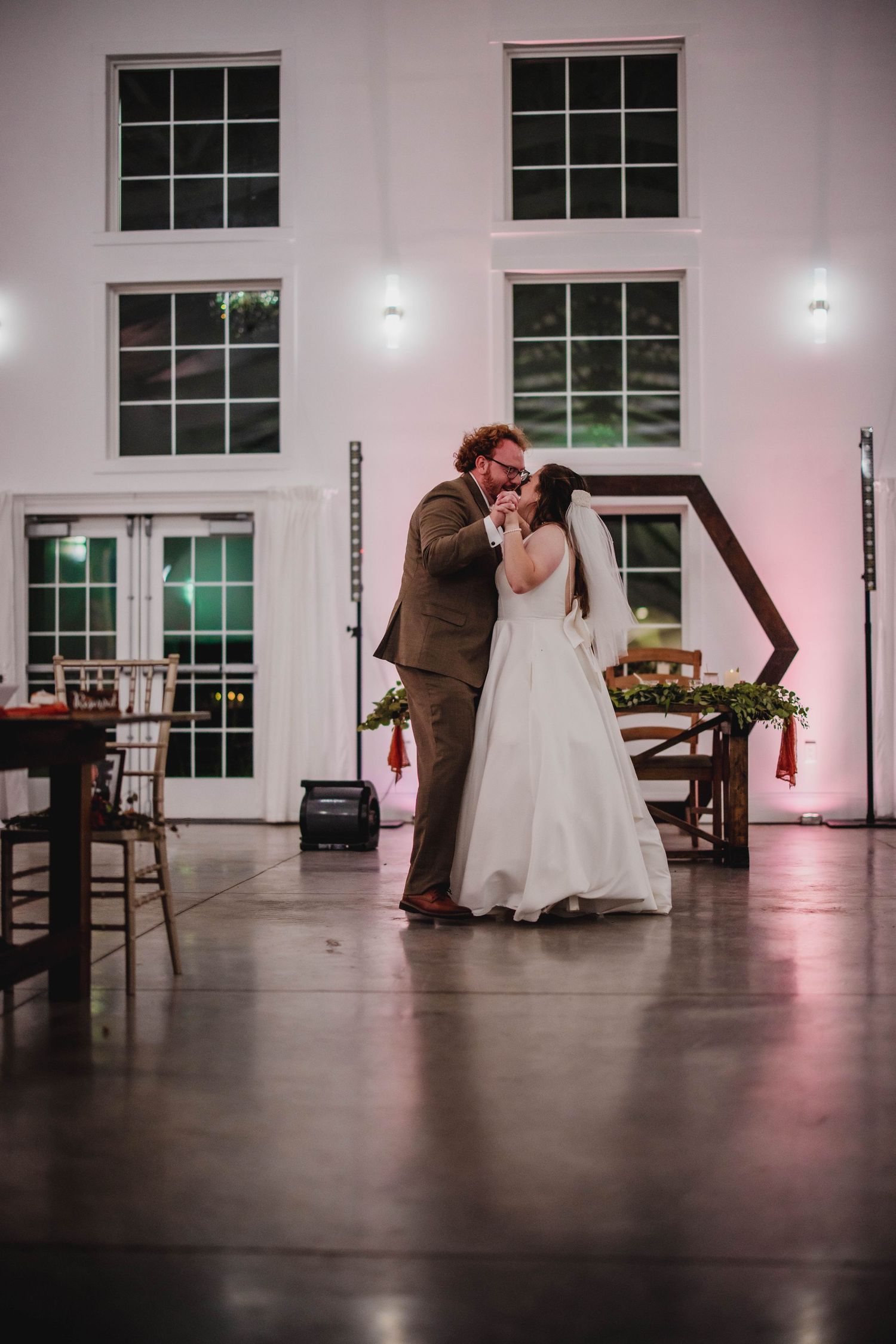 Newlyweds share an intimate dance moment in a modern venue with large windows and pink lighting.
