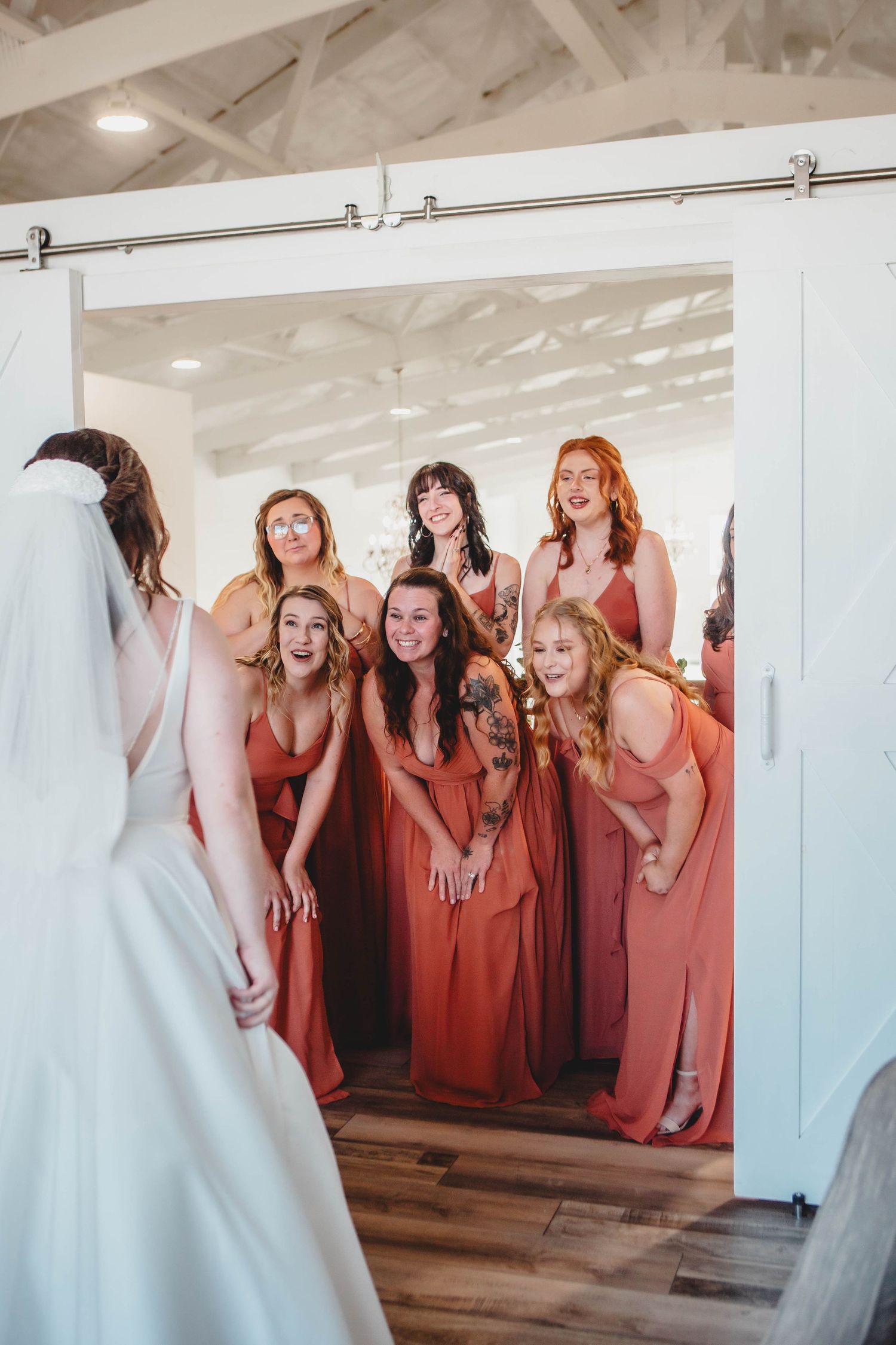 Bride in white gown reveals herself to bridesmaids wearing coral dresses through sliding barn door.