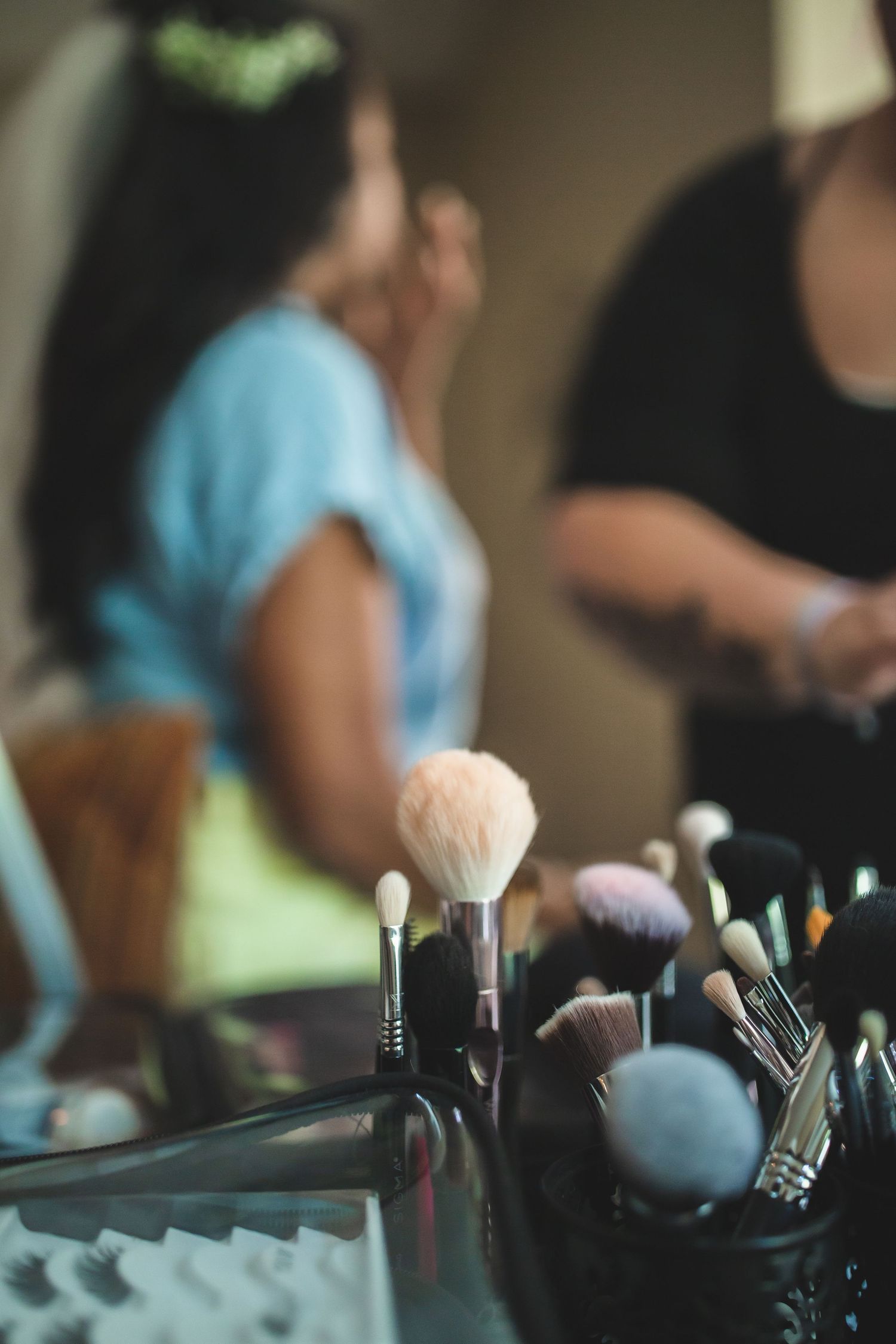 Makeup brushes in foreground with bridal preparation scene blurred in background.