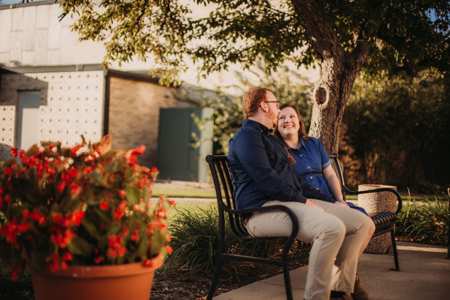 Couple relaxing on park bench surrounded by red flowers and greenery.