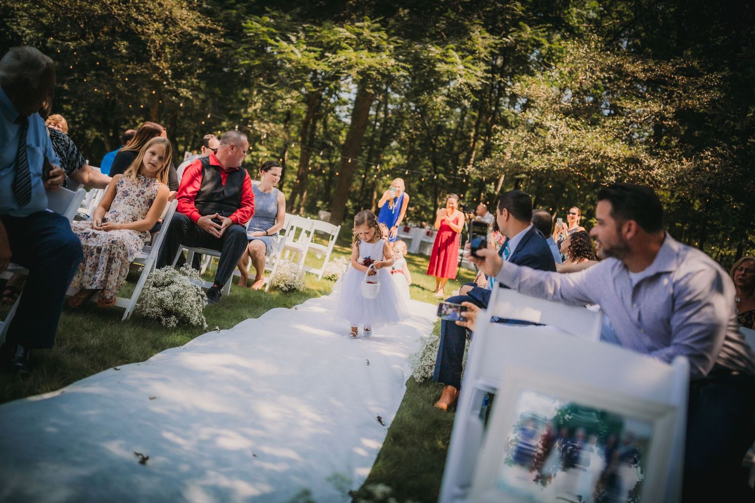 A young attendant walks down an outdoor aisle runner while guests watch from white chairs.