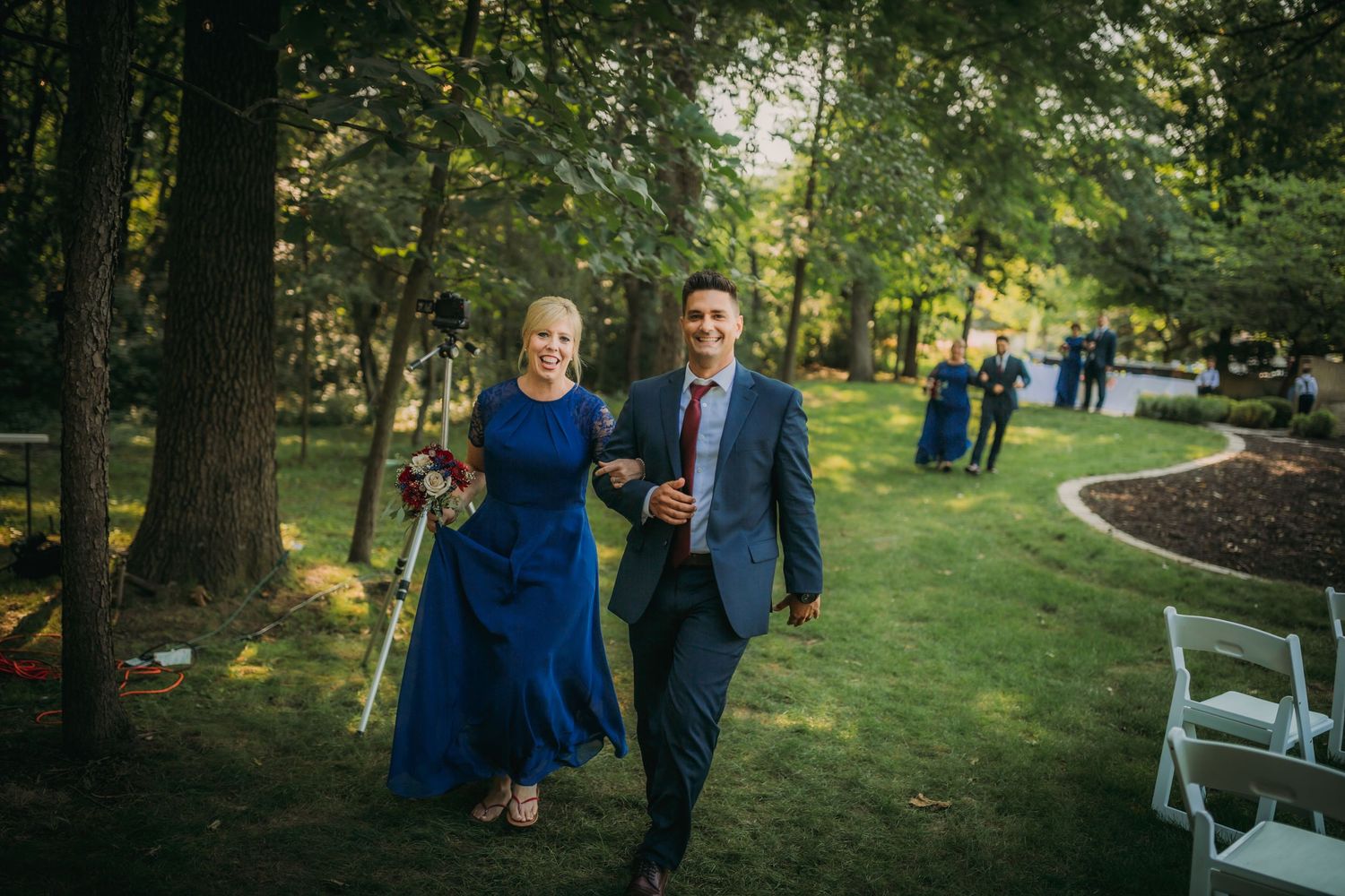 A pair of wedding party members walk arm in arm down a garden ceremony aisle.