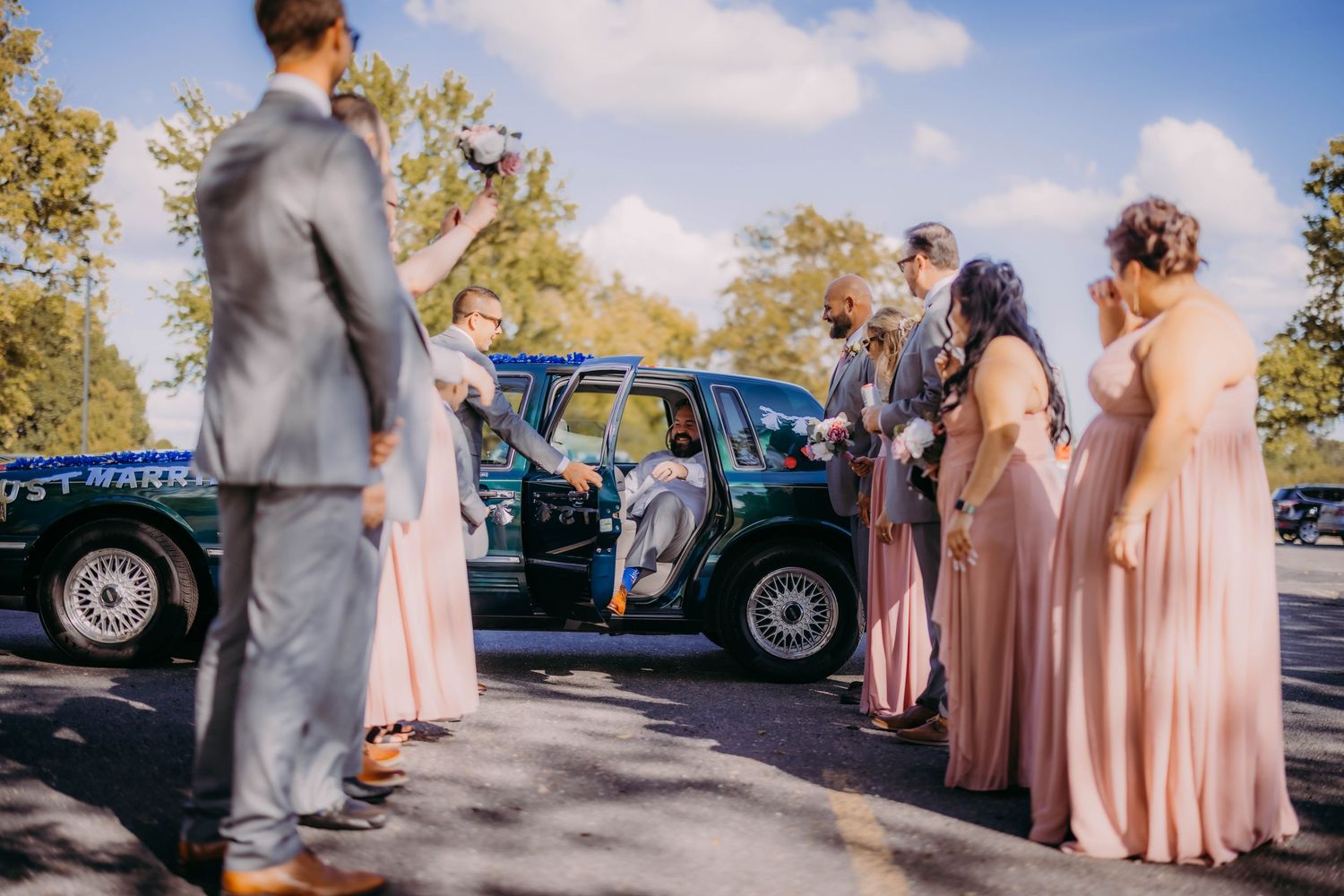 Bridal party in soft pink dresses and grey suits mingles near parked vintage cars.
