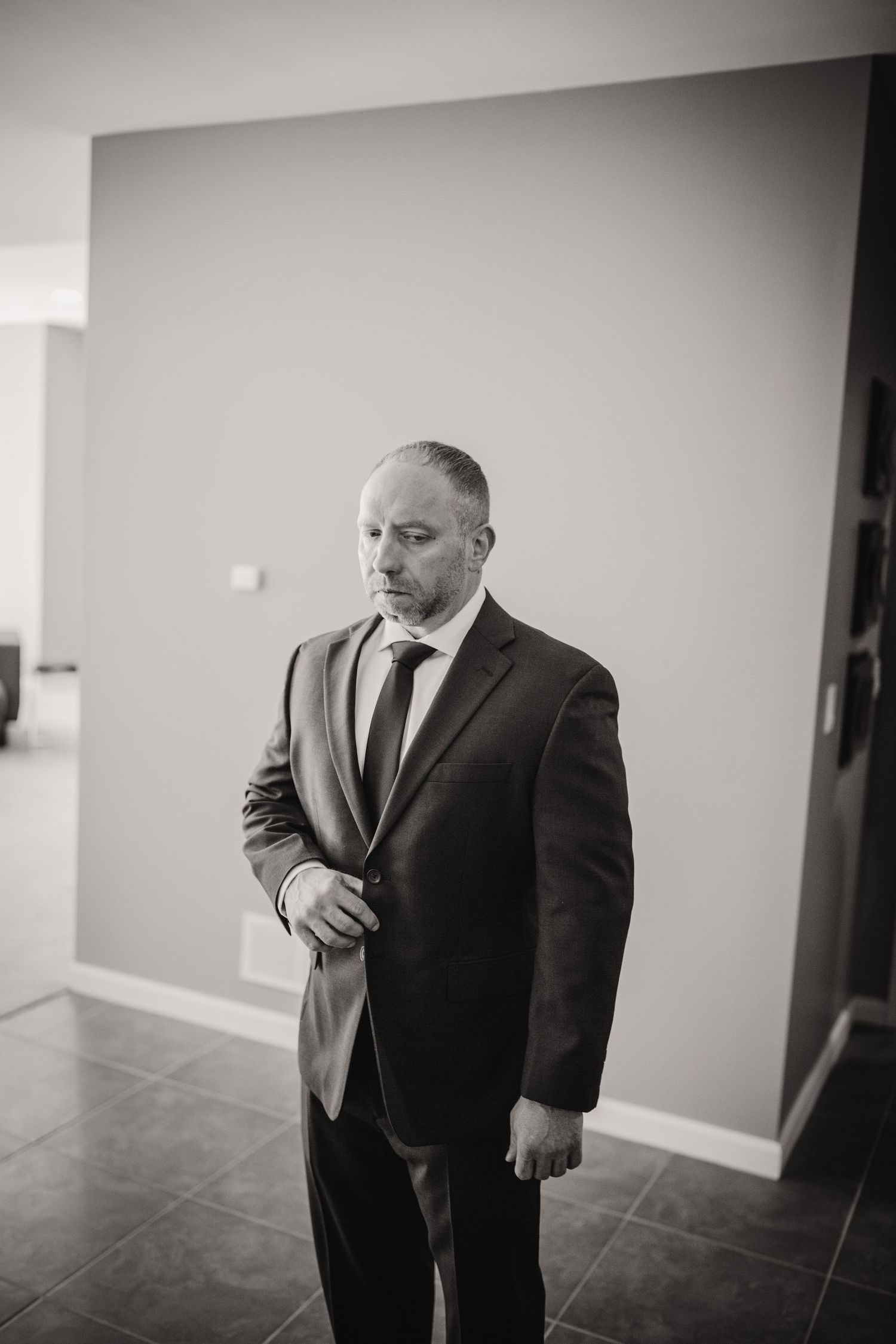 Man in dark suit adjusts his jacket while standing in an empty room with minimal decor.