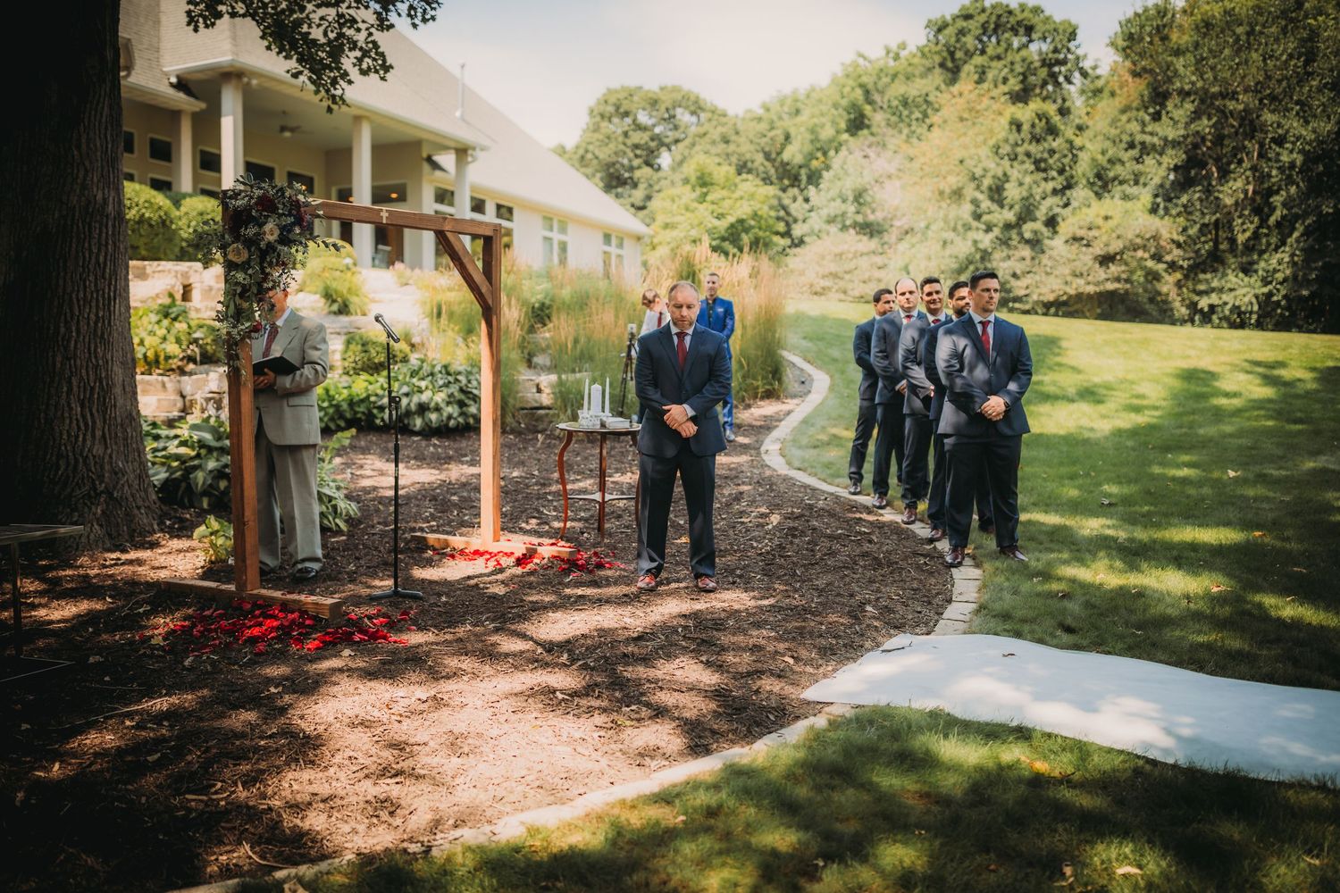 Wedding party members stand along a curved garden pathway during ceremony preparations.