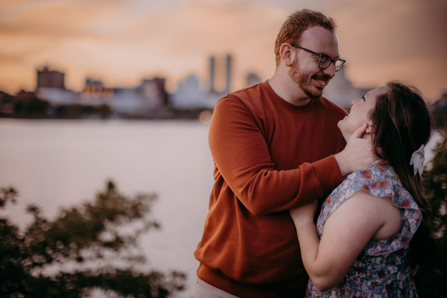 A couple shares an intimate moment at sunset with a city skyline in the background on the waterfront.