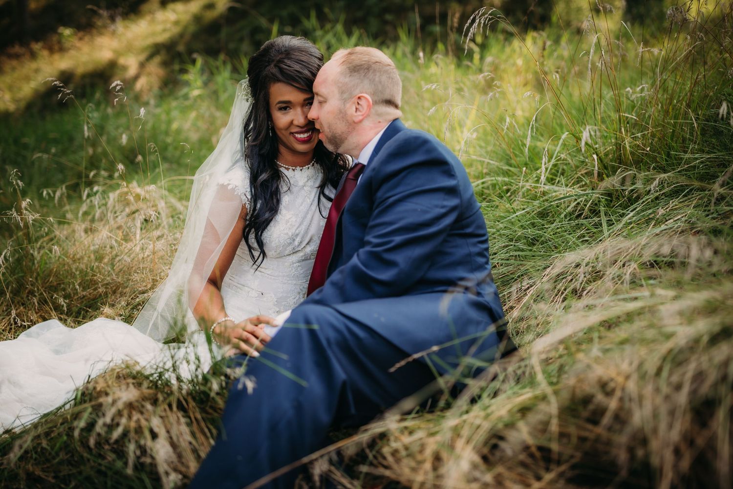 Joyful wedding moment captured in rustic outdoor setting with couple sitting among tall grasses.