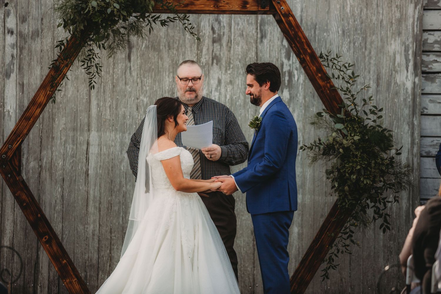 Outdoor wedding ceremony features wooden triangle arch decorated with greenery against weathered barn wall.
