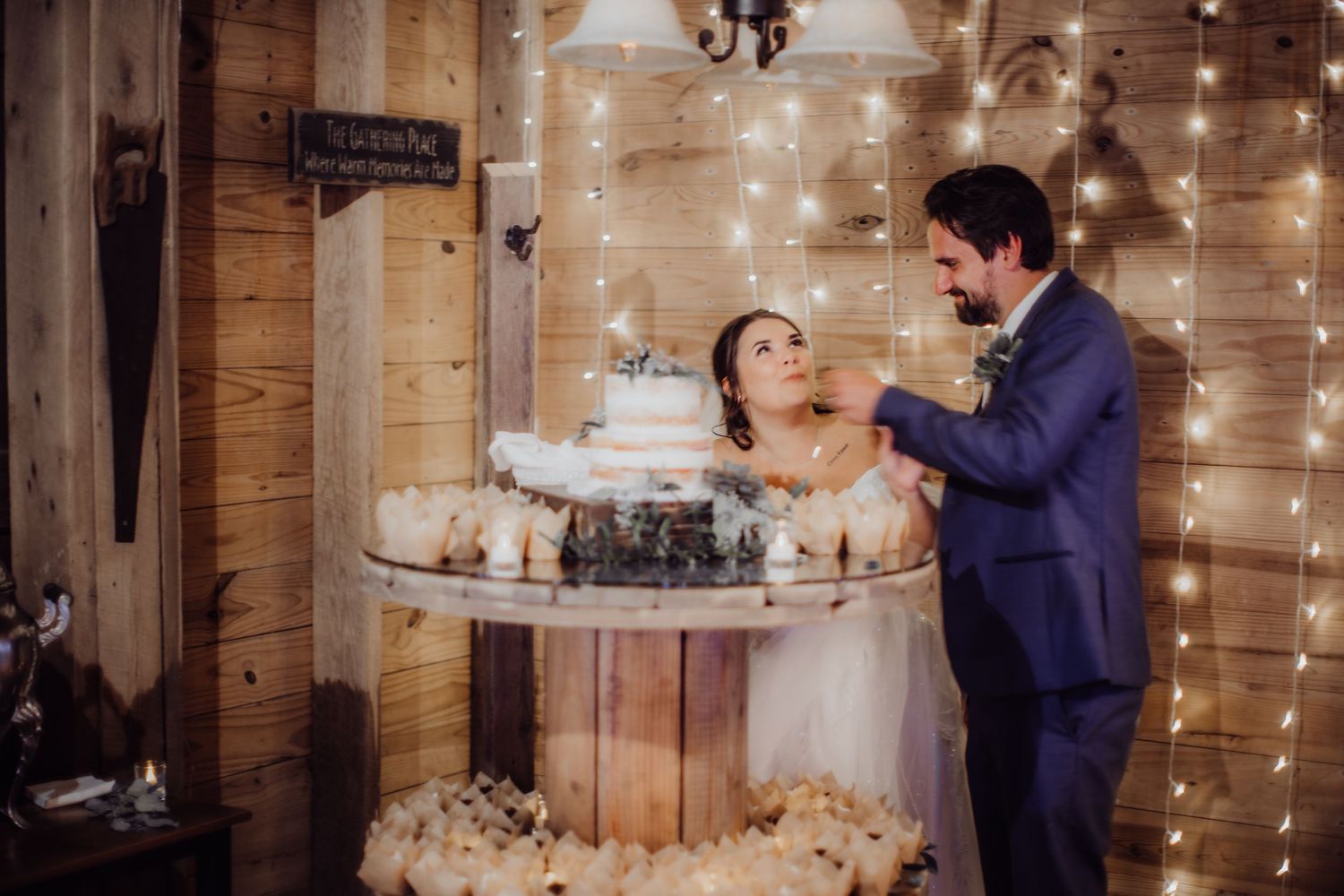 Couple feeding each other wedding cake surrounded by twinkle lights in a rustic barn setting.