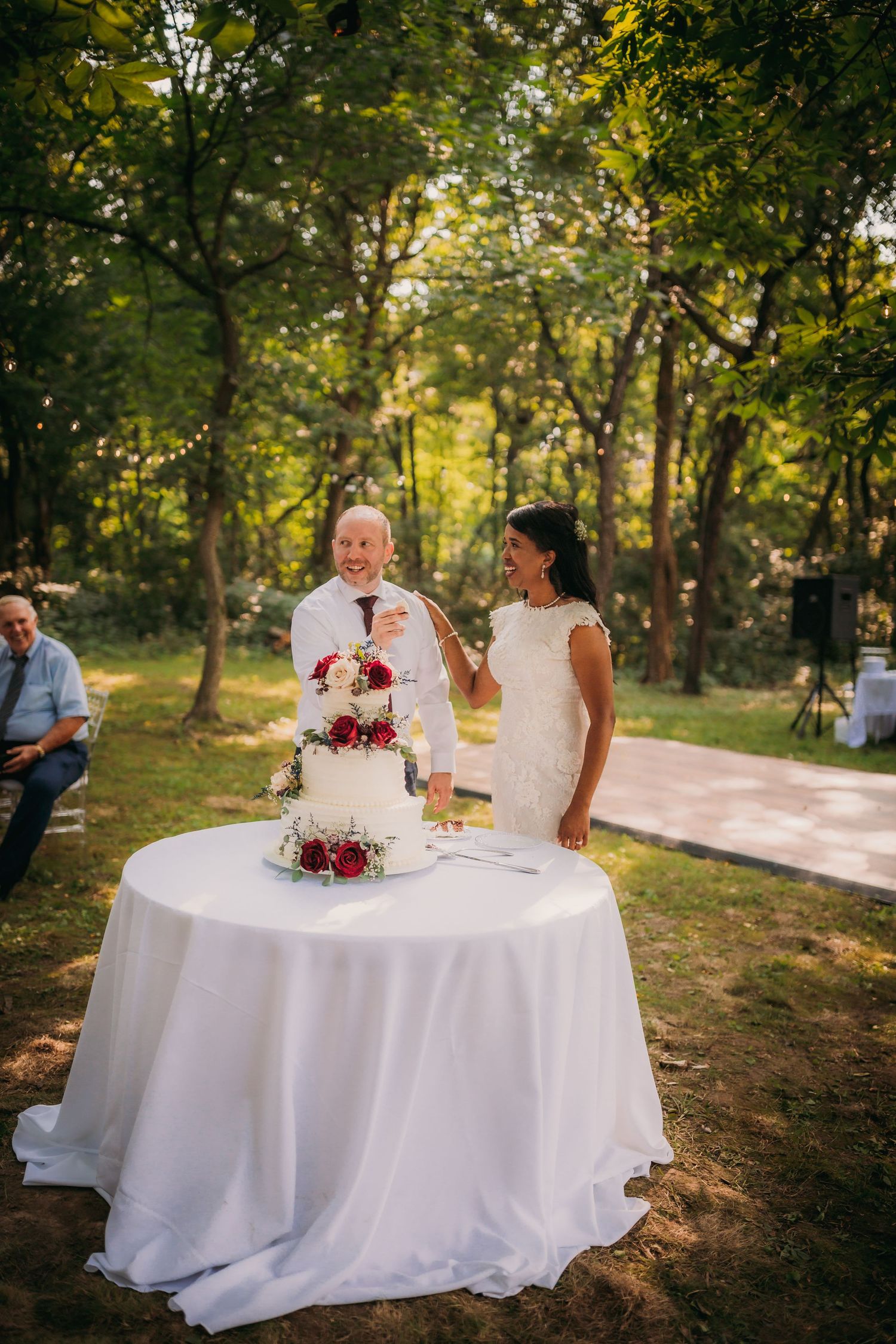 Outdoor wedding ceremony with white table and elegant cake against a backdrop of trees and string lights.