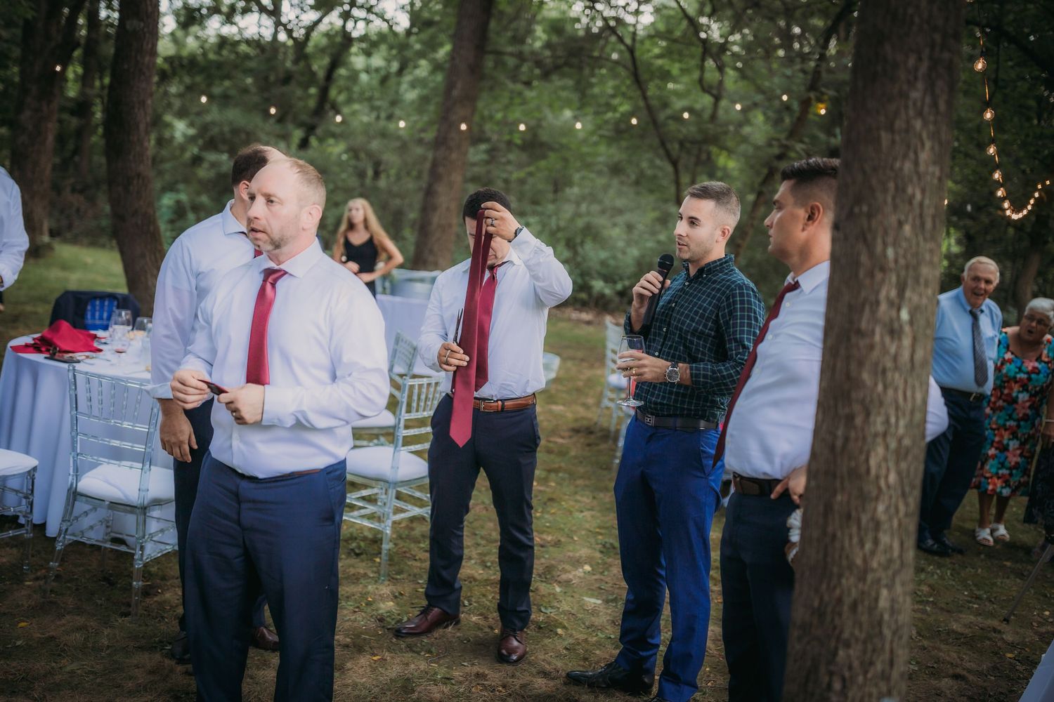 Guests in formal attire gather at an outdoor wedding reception with string lights and white chairs.