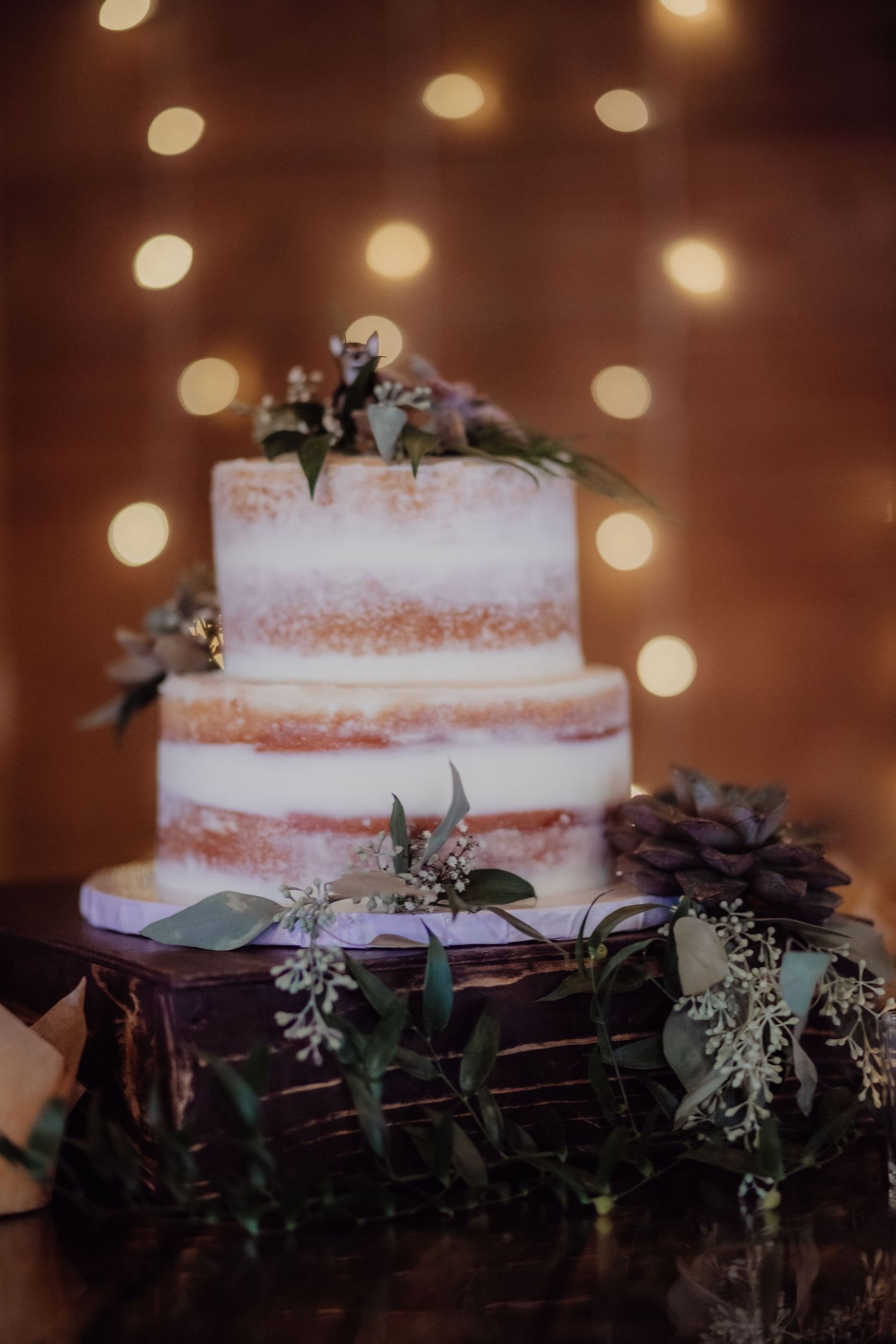 Semi-naked wedding cake with white frosting and floral decorations against bokeh lights.
