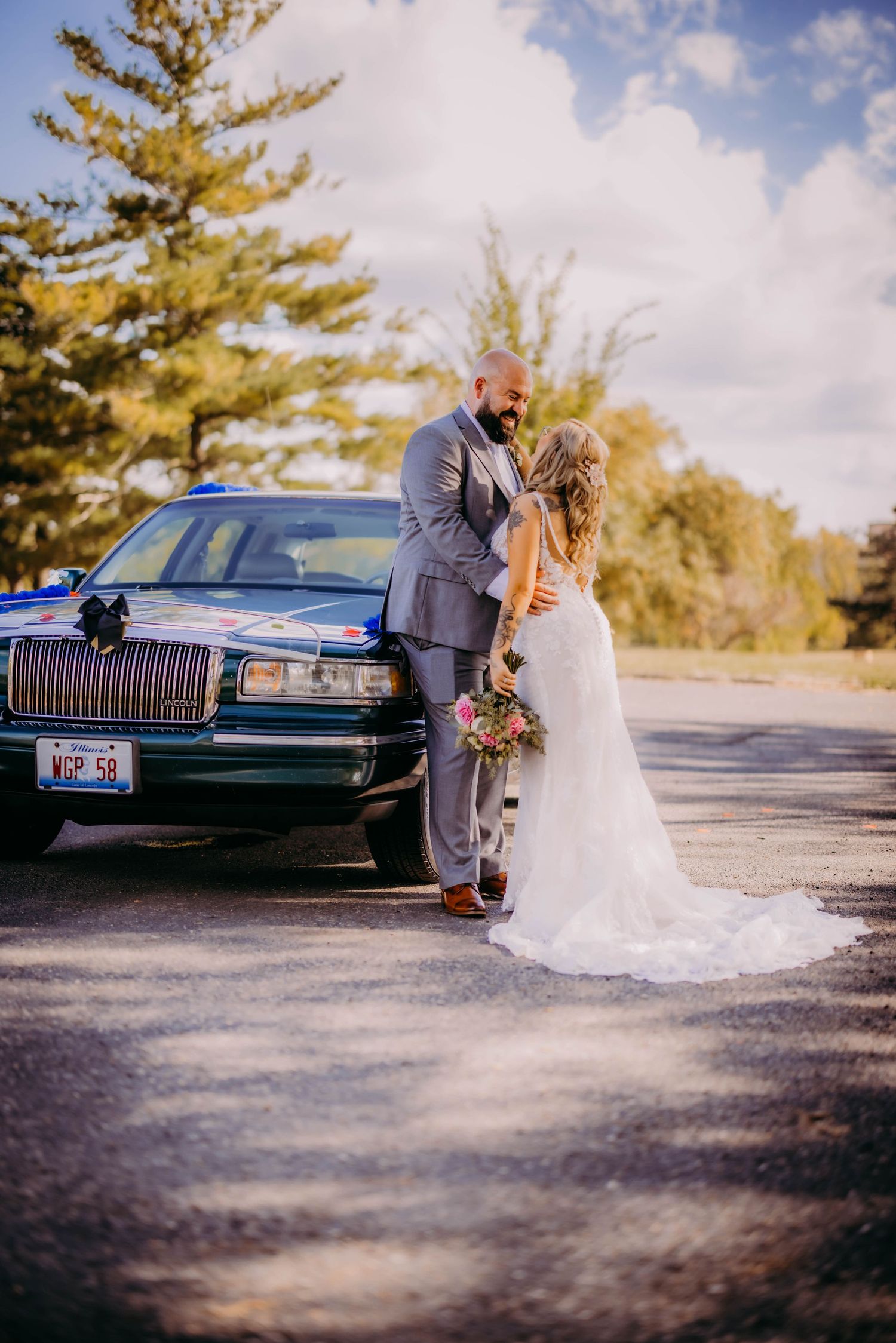 Wedding couple poses with vintage Lincoln car outdoors against autumn trees and cloudy sky.