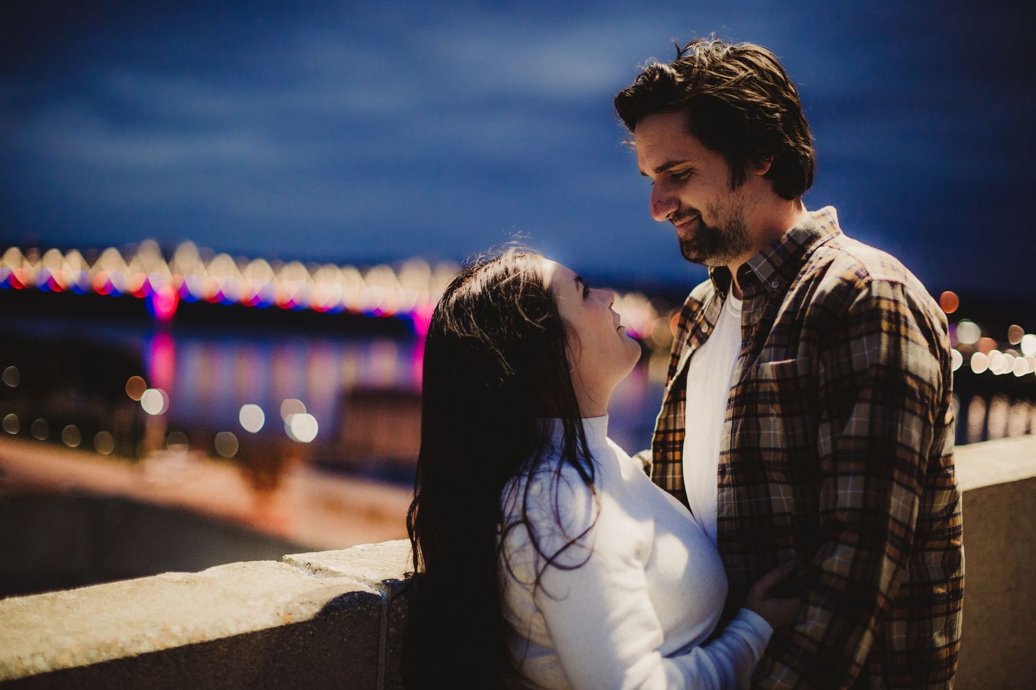 A couple shares an intimate moment outdoors at dusk with city lights glowing in the distance.