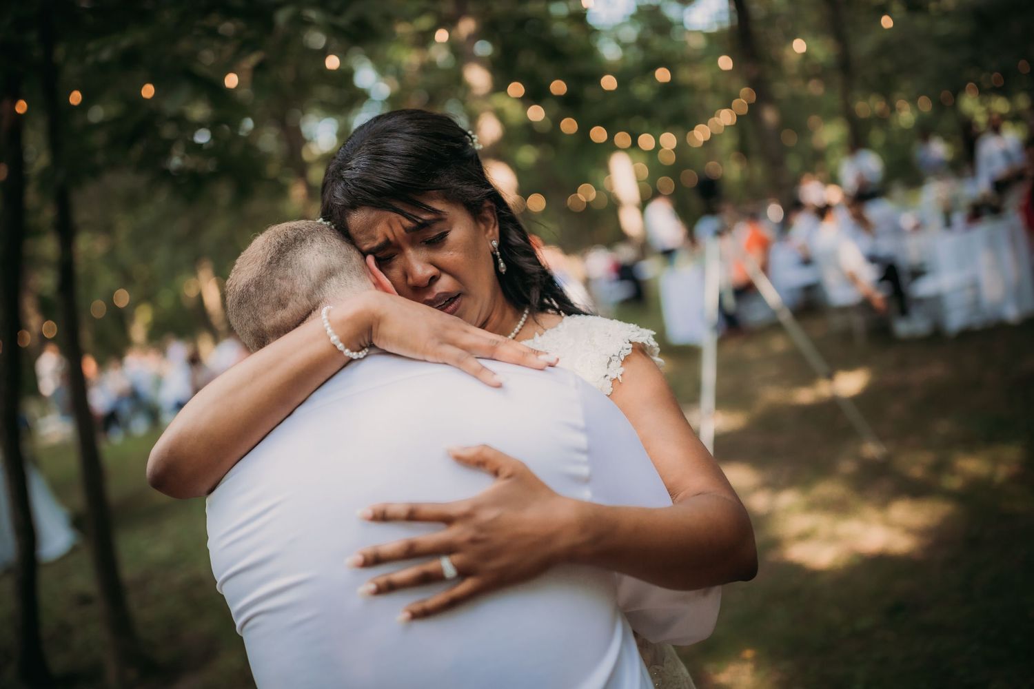 Close embrace during wedding dance captured in scenic outdoor wooded setting with string lights overhead.