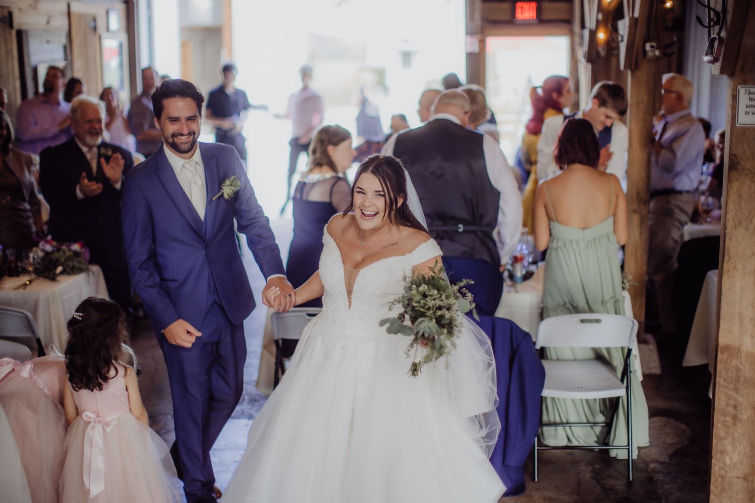 Joyful couple walks through crowd of wedding guests in rustic indoor venue with natural lighting.