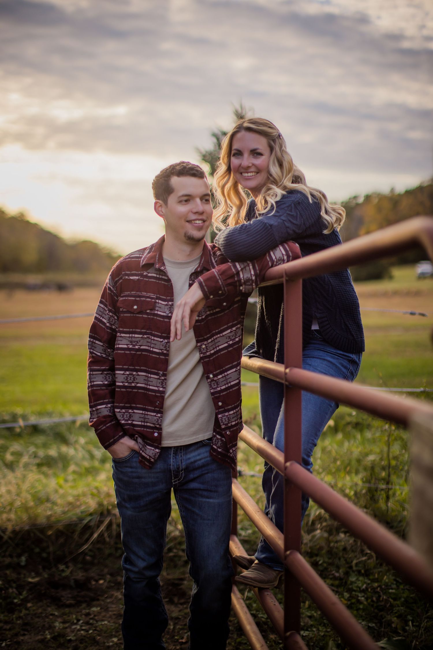 Casual couple leans against rustic fence with golden fall scenery in background.