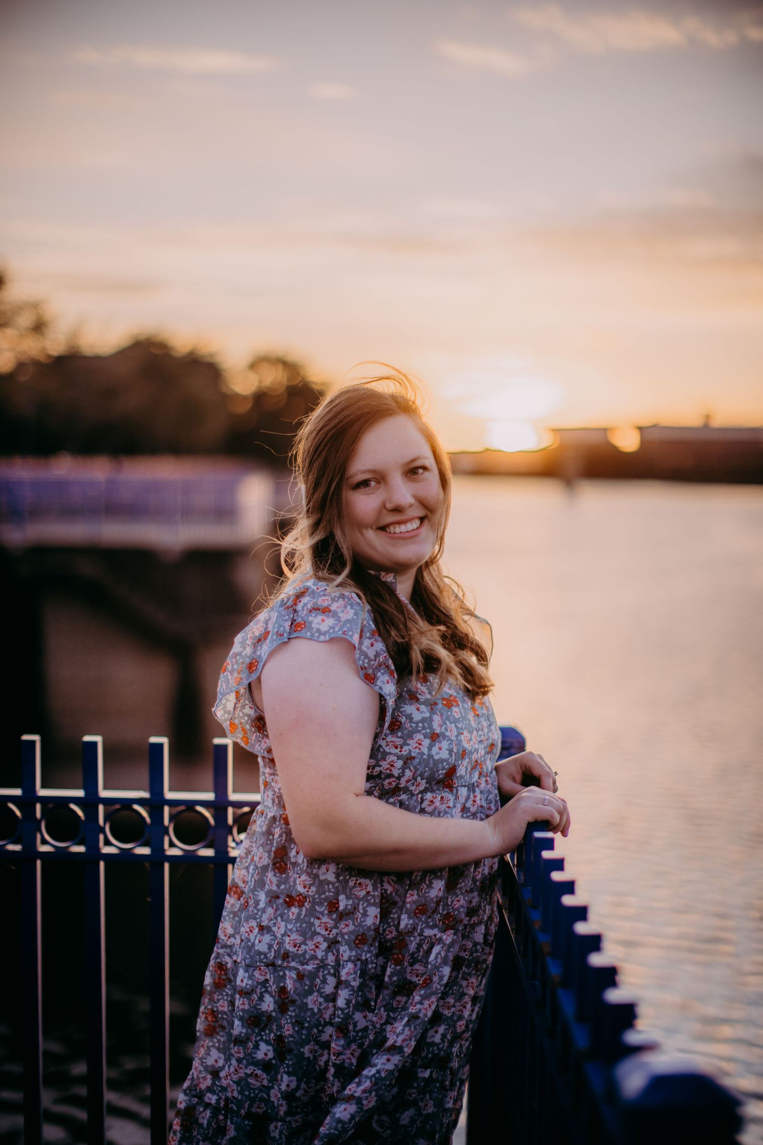 Woman in patterned dress smiles warmly by waterfront during sunset.