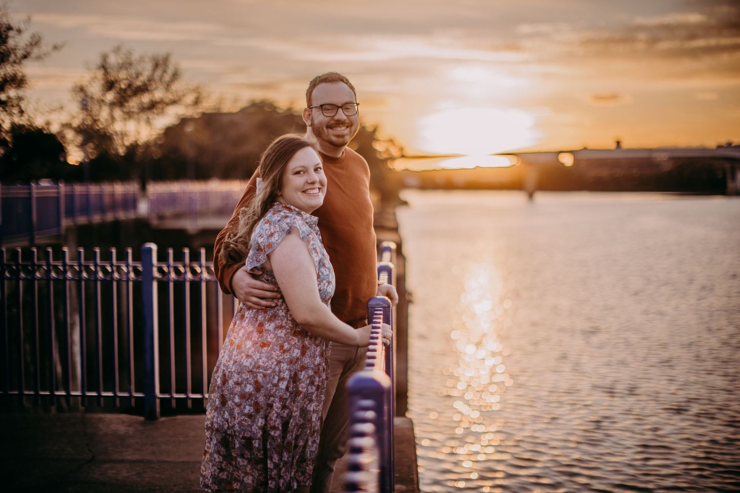Golden hour portrait session along waterfront with metal railings and glowing sunset.
