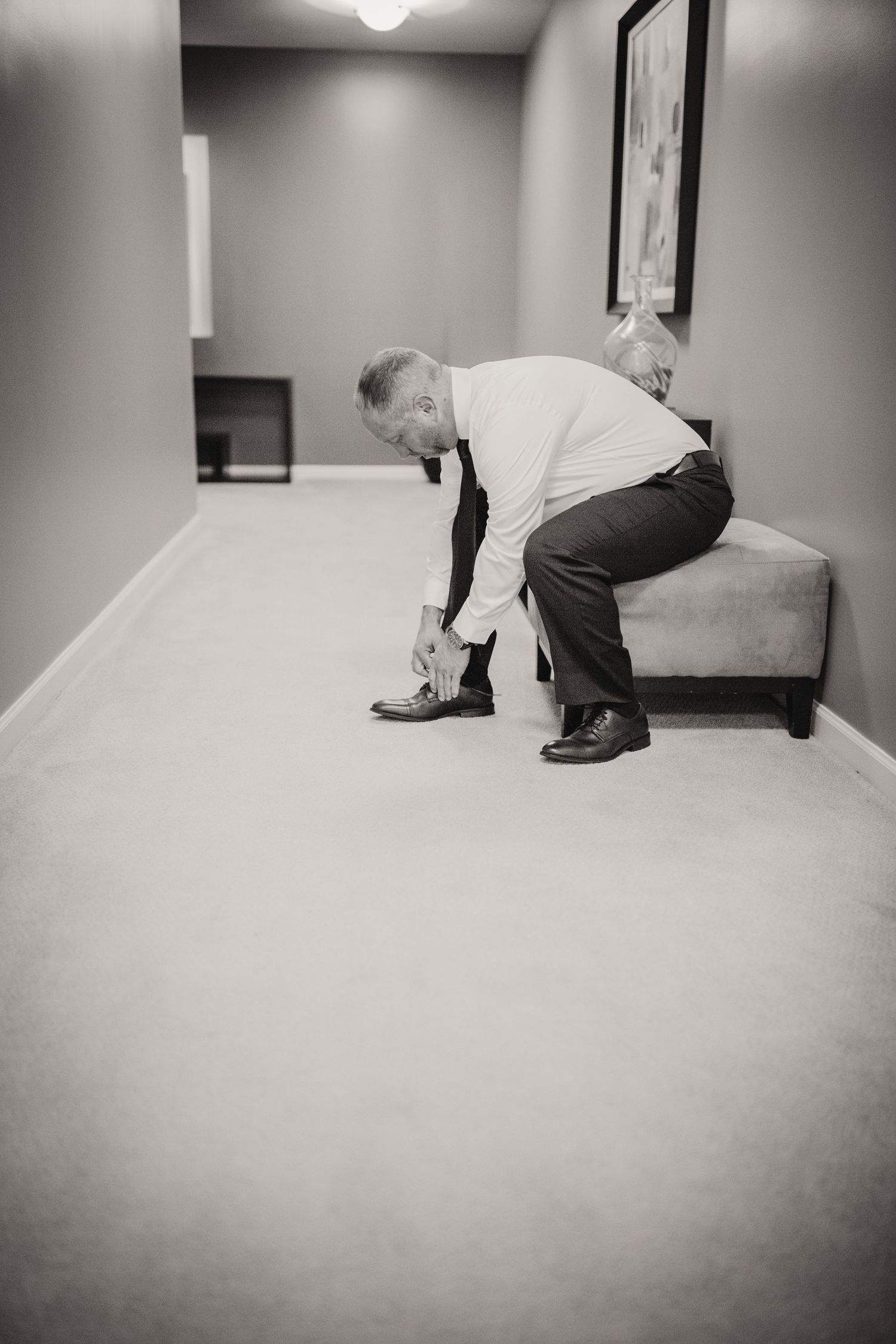 A black and white photo shows someone tying their dress shoes in a carpeted hallway.