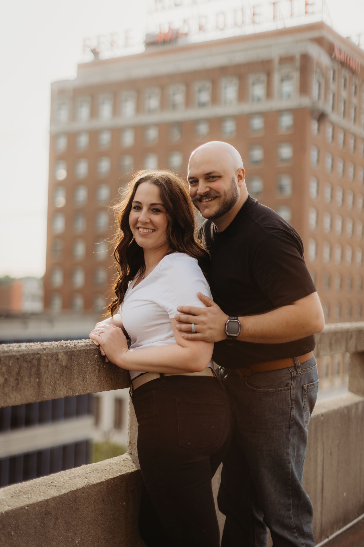 A couple poses together on an urban rooftop with the Marquette Hotel building in the background.