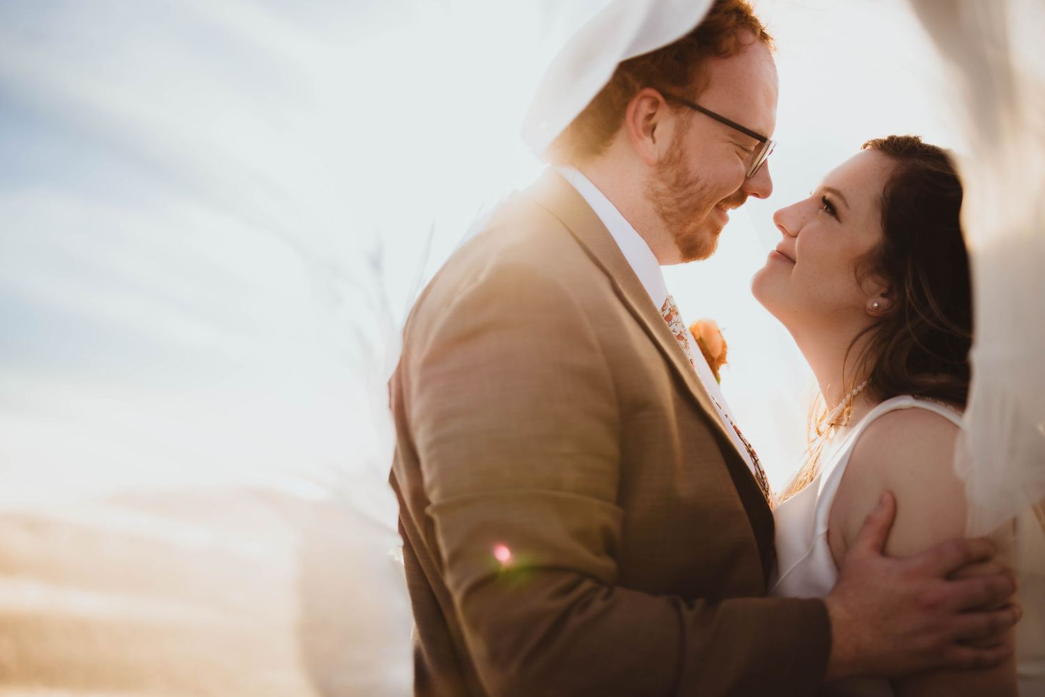 Wedding couple embracing in soft natural light with a delicate veil surrounding them.