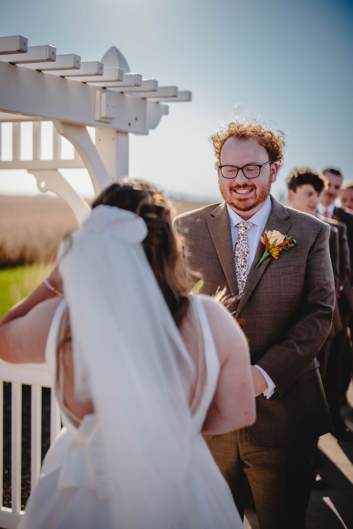 Sunny outdoor wedding ceremony with white pergola and floral boutonniere detail.