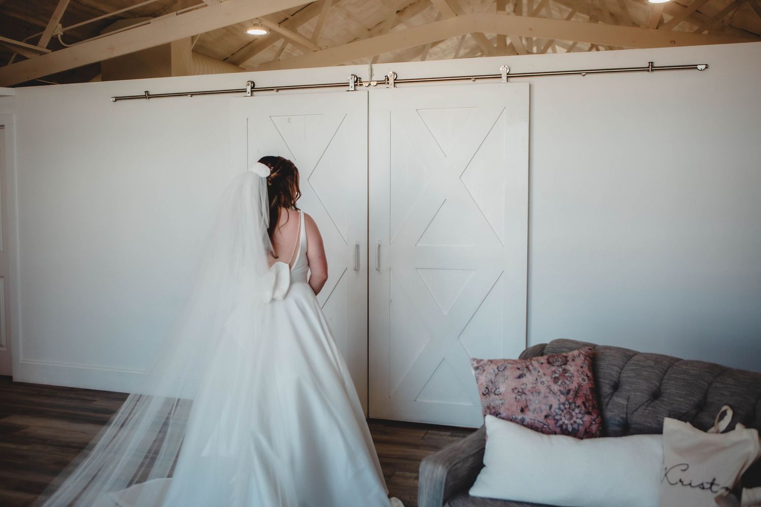 Bride in flowing white gown stands before modern sliding barn door in rustic venue.
