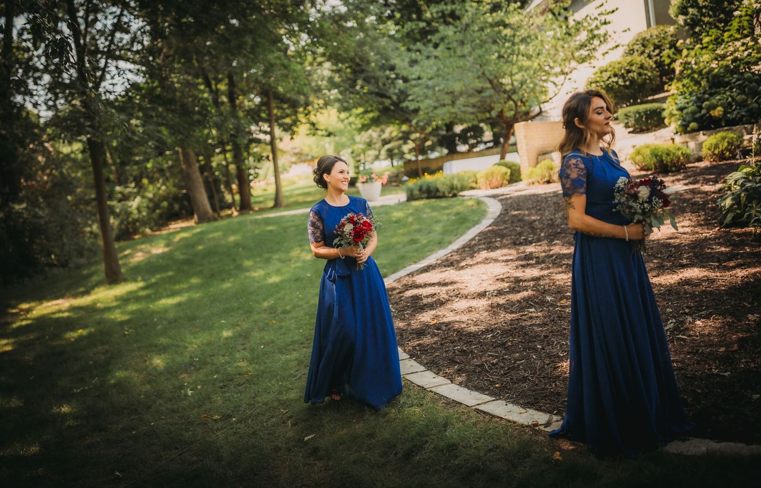 Bridesmaids in royal blue dresses carry bouquets while walking through garden setting.