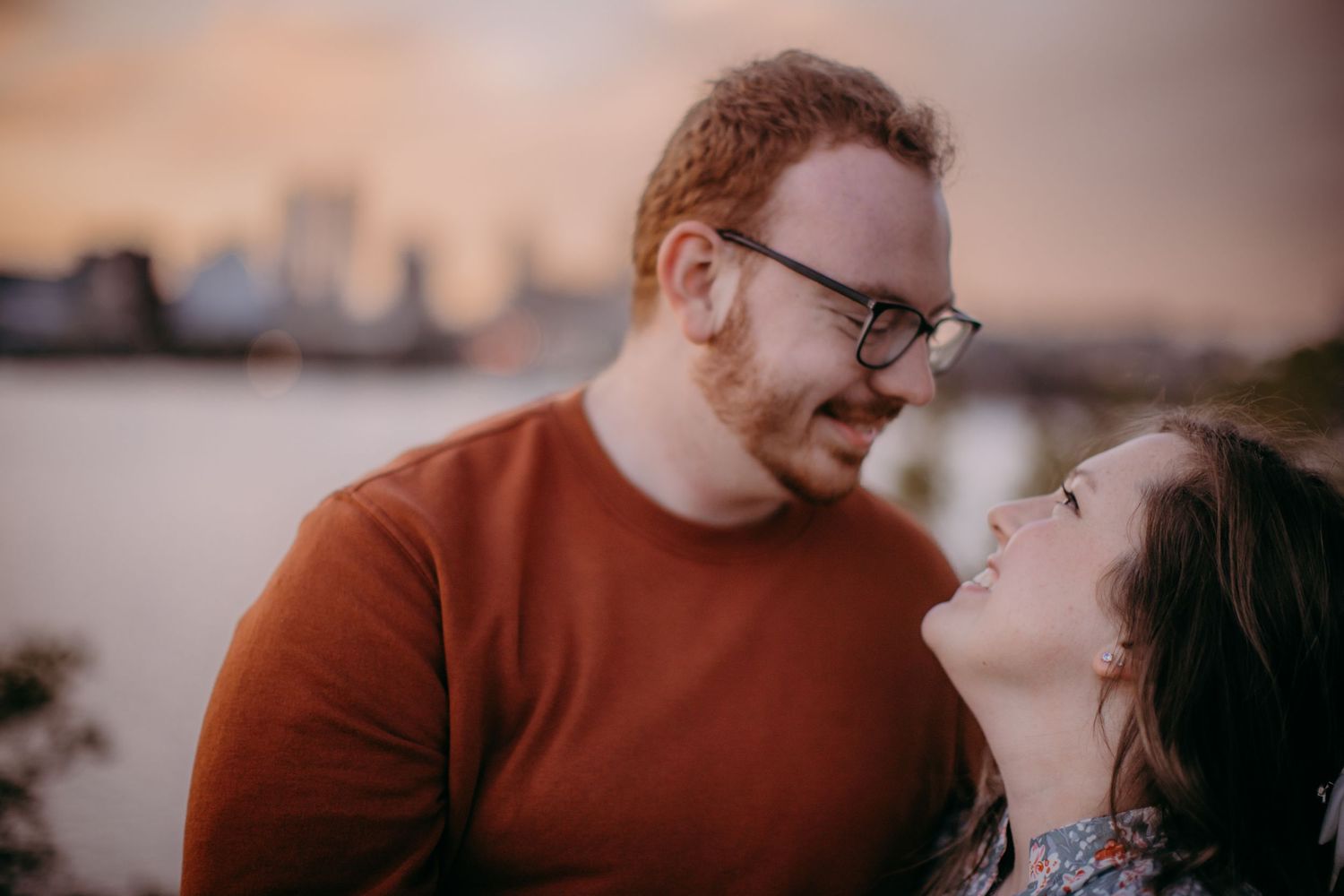 A tender moment between a couple against a blurred waterfront cityscape at dusk.