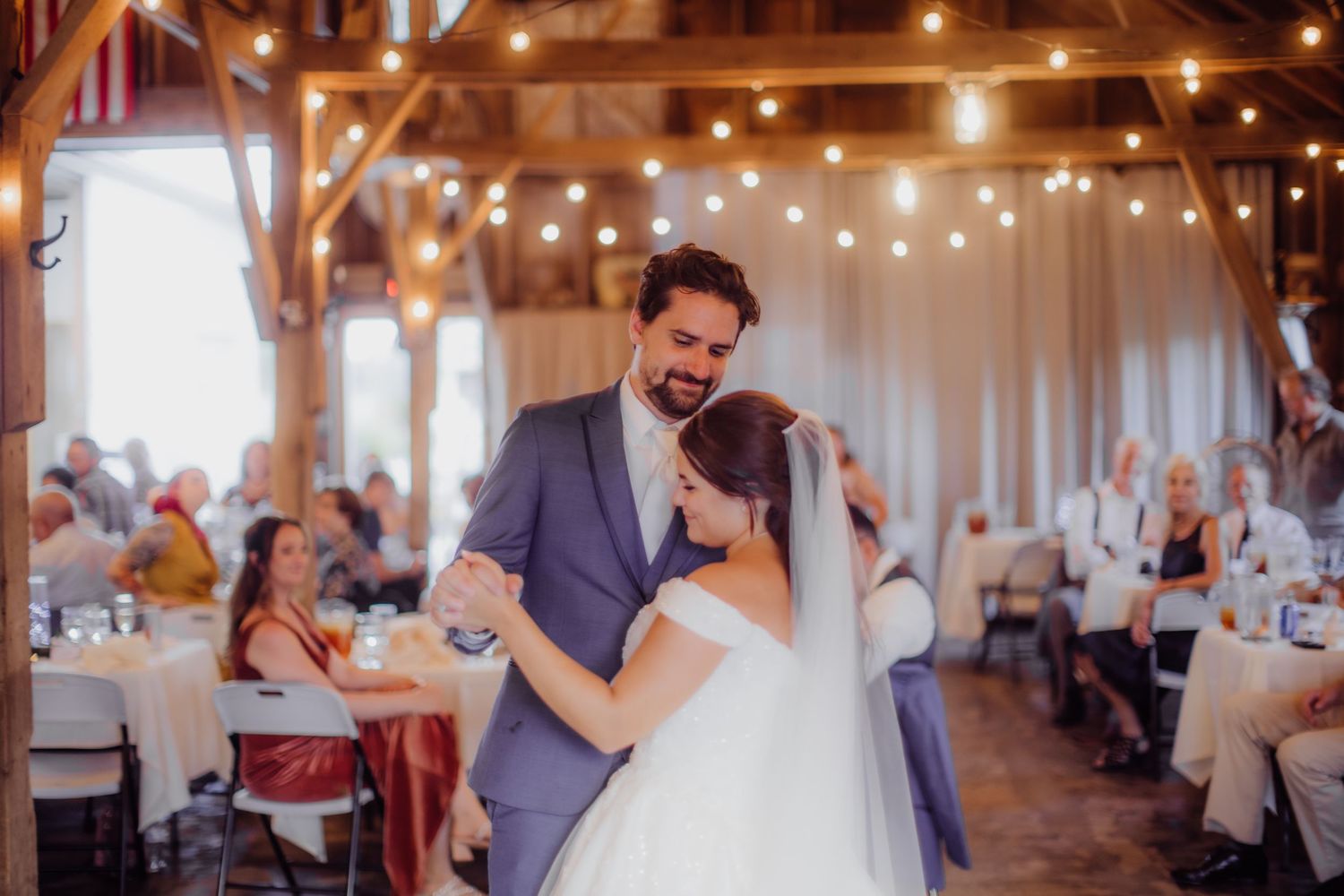 Couple sharing their first dance in a rustic barn venue illuminated by string lights.