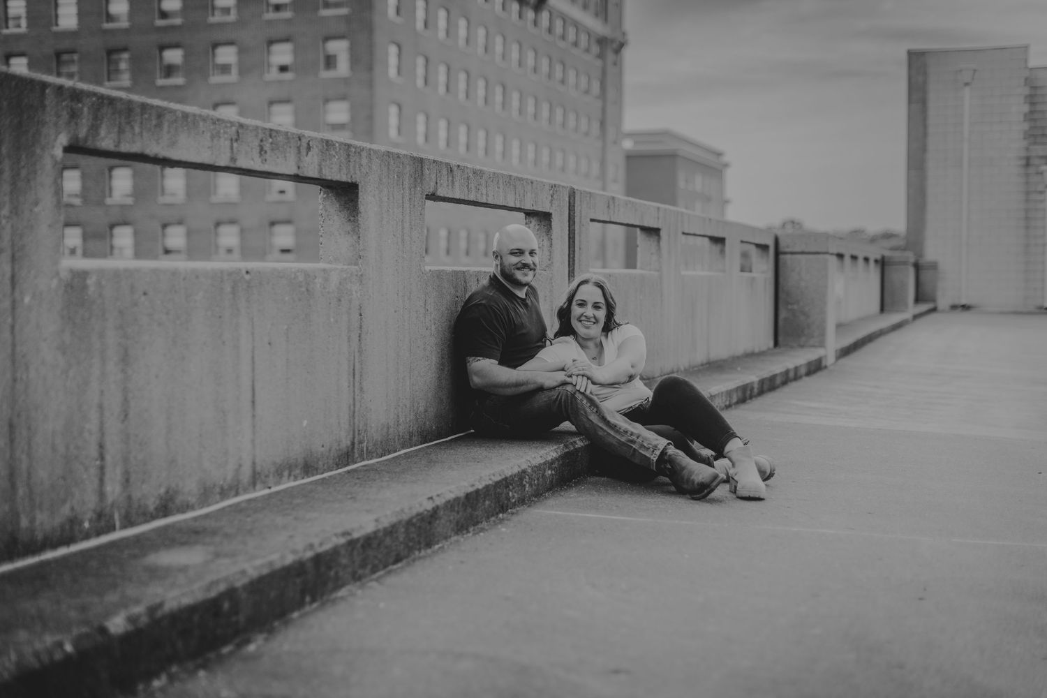 A couple sits together on a concrete ledge against an urban backdrop of brick buildings in black and white.